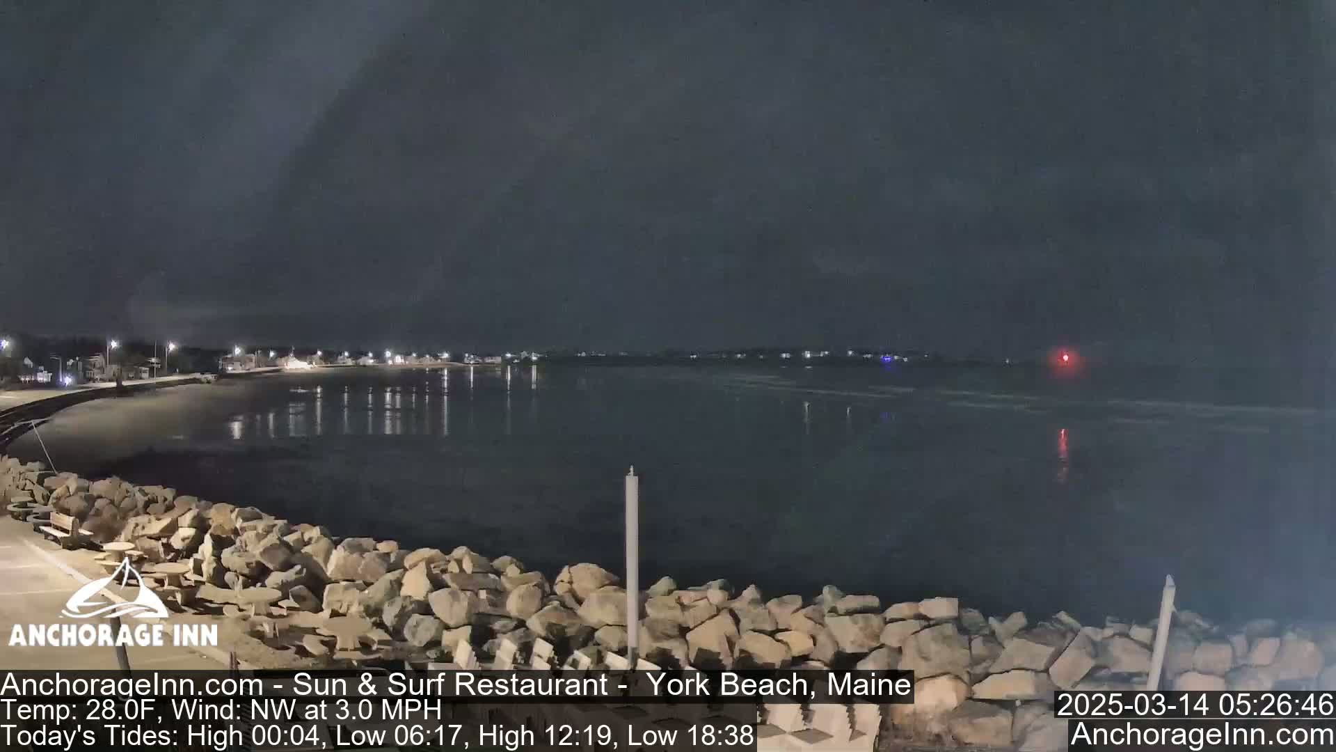 A nighttime view of a calm, dark body of water next to a rocky shoreline, with a few lights visible in the distance under a mostly clear sky.