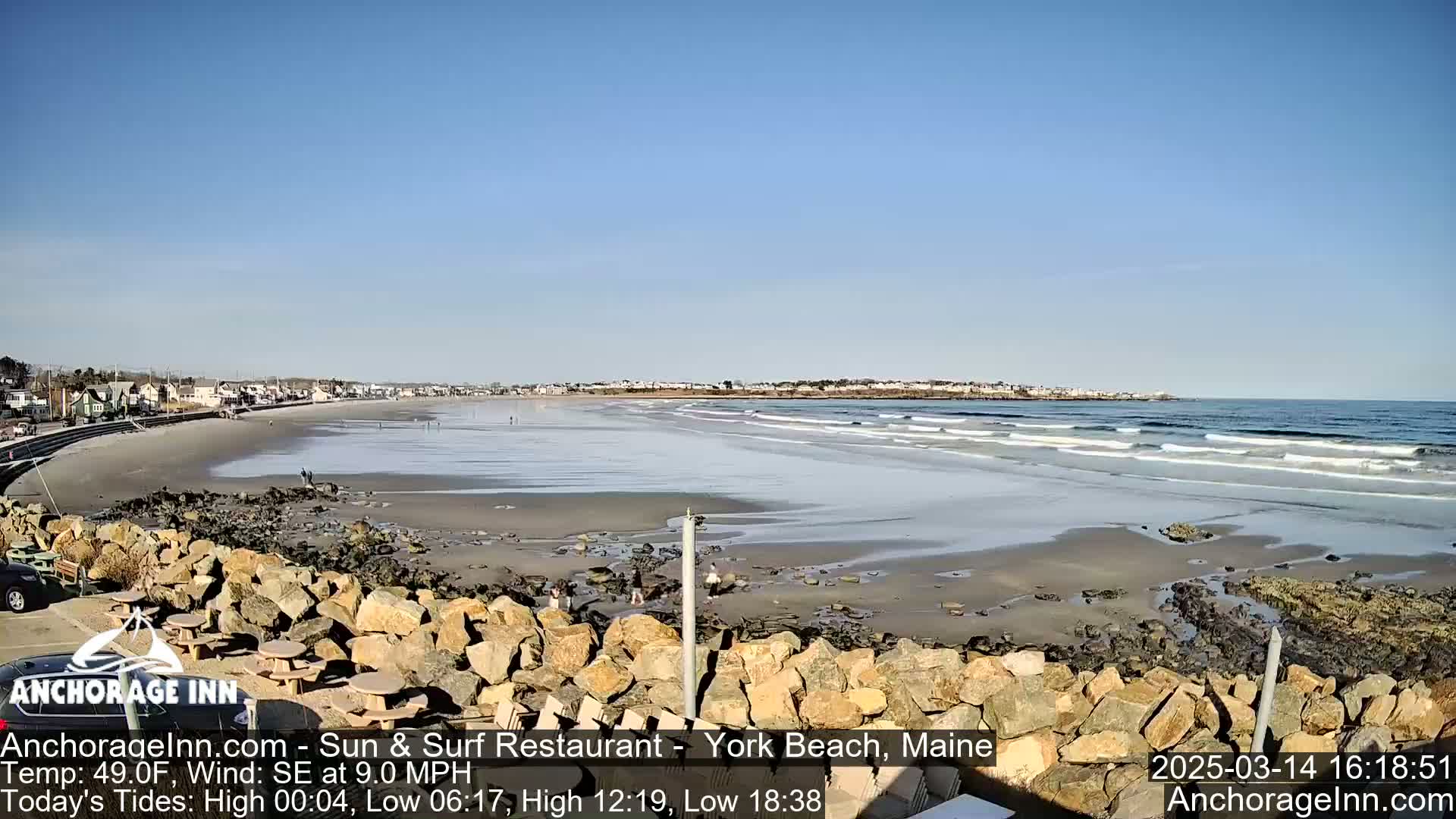 A sandy beach with gentle waves under a clear blue sky is viewed from behind a low rock wall.