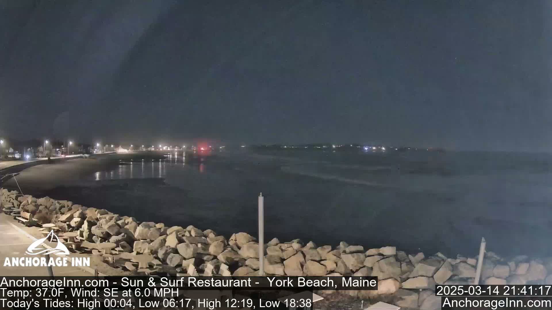 A dark, nighttime scene shows a rocky shoreline and calm ocean water under a clear sky, with distant lights visible across the water.
