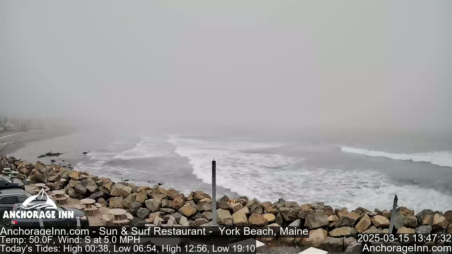 A foggy beach scene shows a rocky shoreline with small waves lapping against it.