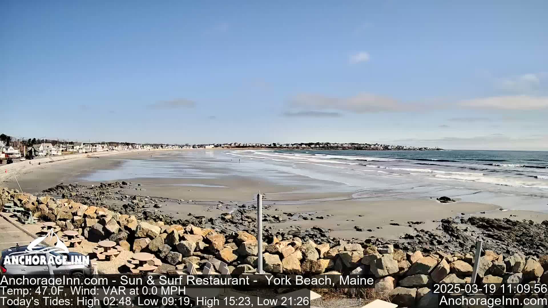 A sandy beach with a calm ocean and a clear blue sky is seen from a rocky vantage point.
