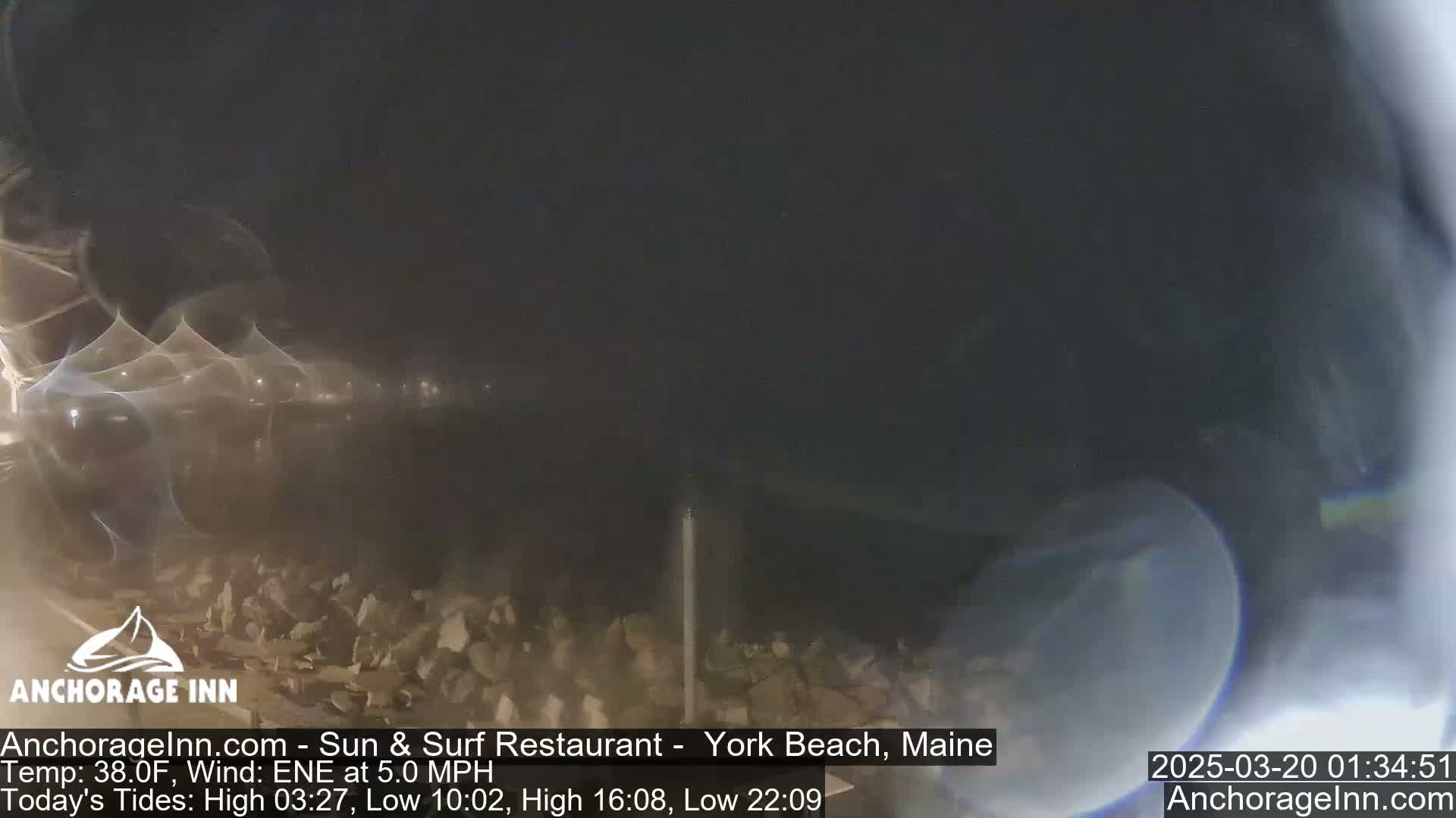 A nighttime, blurry view of a rocky shoreline under a dark sky with a light east-northeast wind.