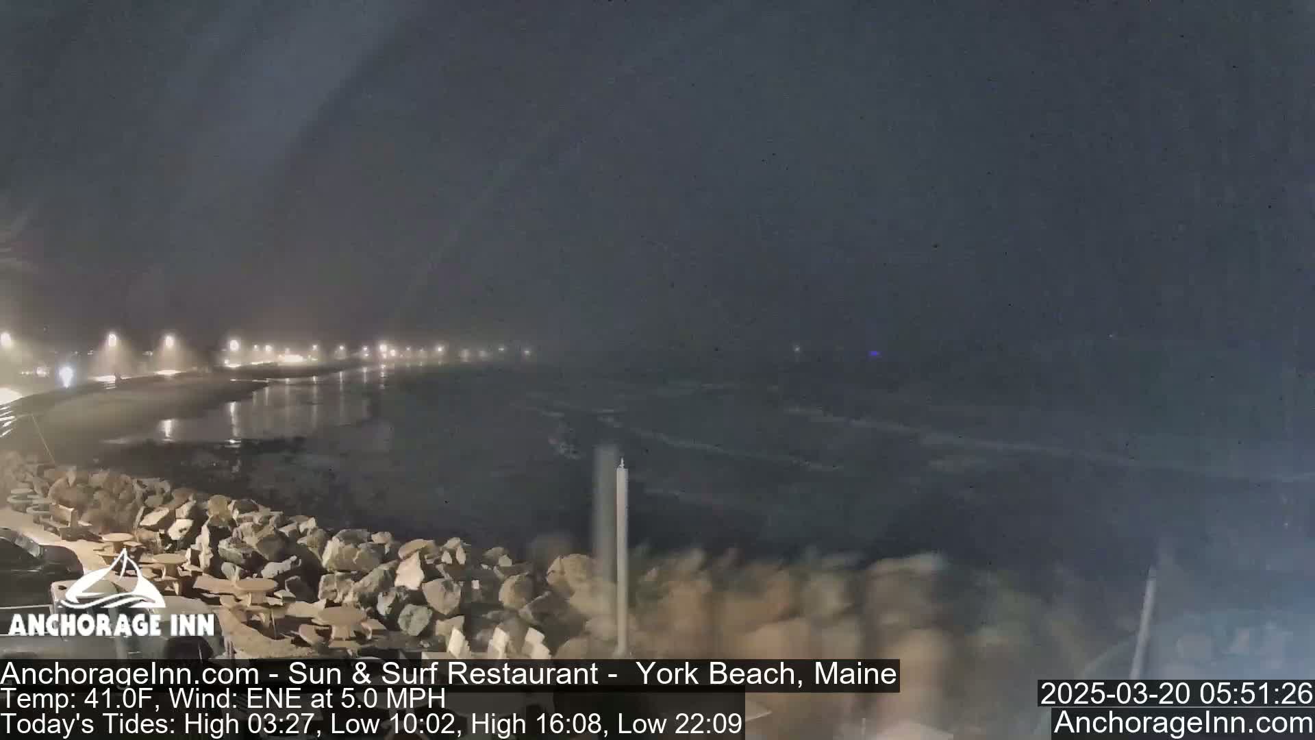 A nighttime coastal scene shows a dark, somewhat hazy sky over a rocky shoreline and calm, dark water with lights visible in the distance.