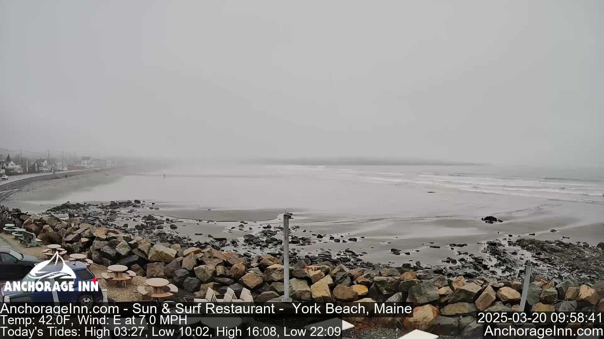 A foggy beach scene shows a rocky shoreline with a calm ocean and a distant road with buildings under an overcast sky.