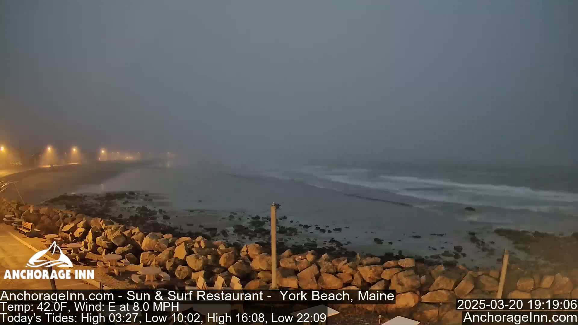 A foggy, dark evening scene shows a rocky shoreline with gentle waves lapping at the beach under a gray sky.