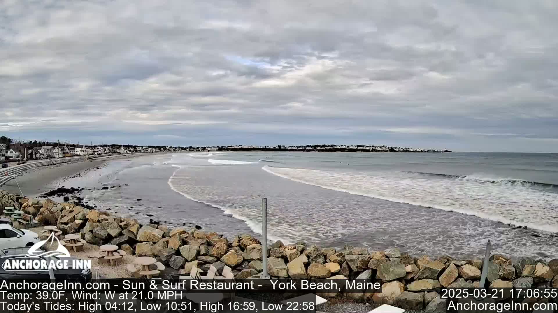 A rocky shoreline overlooks a beach with gentle waves rolling in under a cloudy sky.