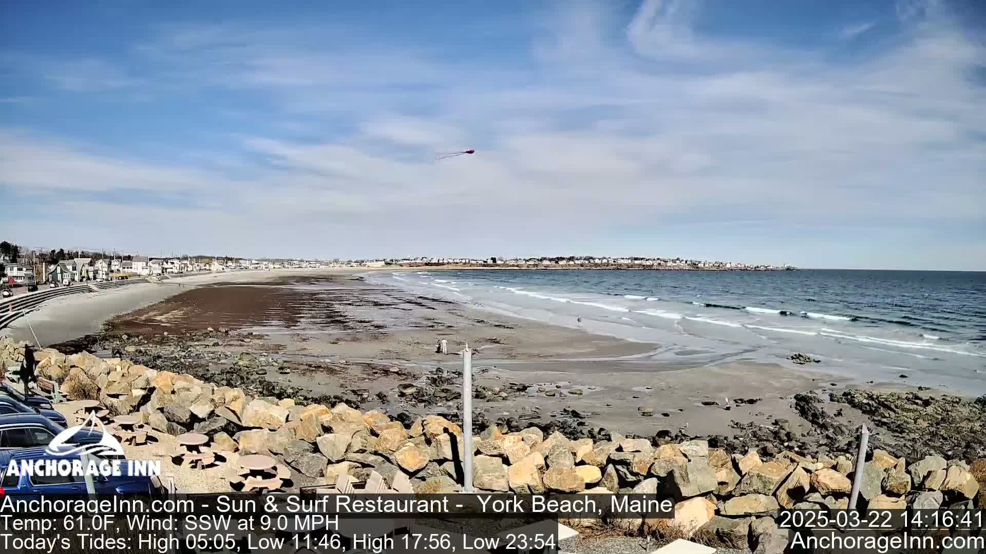 A sandy beach with a rocky shoreline and gentle waves stretches beneath a mostly clear blue sky; a kite is visible in the upper right.