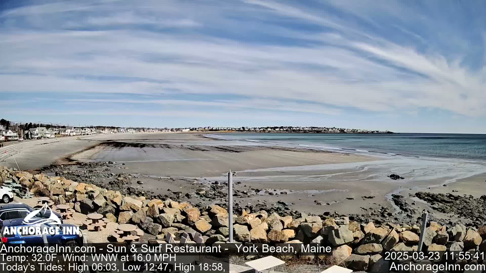 A wide sandy beach stretches to a calm ocean under a mostly clear, bright sky, with a rocky area in the foreground and buildings visible in the distance.
