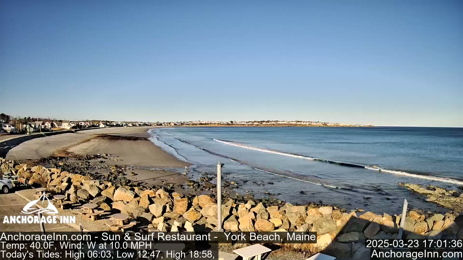 A sandy beach with gentle waves laps the shore under a clear blue sky, backed by a rocky outcrop and a row of houses.