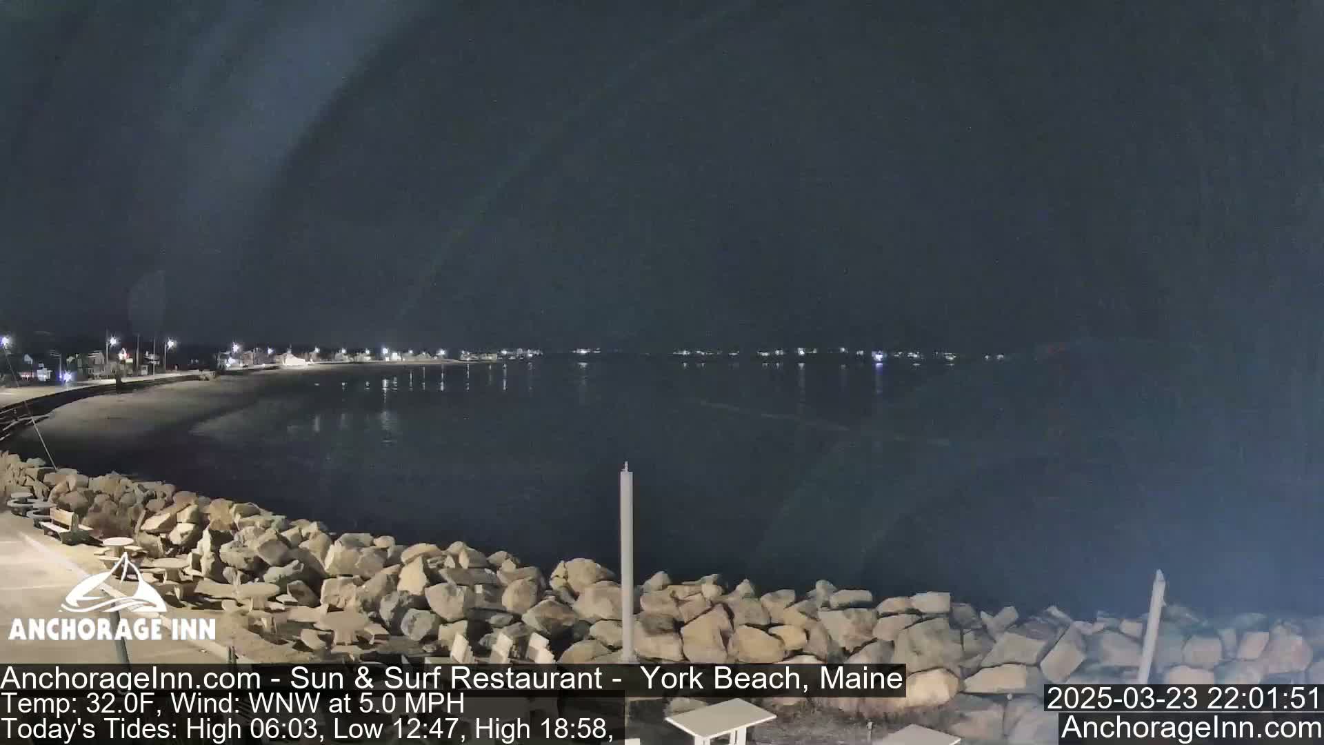A nighttime view of a rocky shoreline and calm water, with a town faintly lit in the distance under a dark sky.