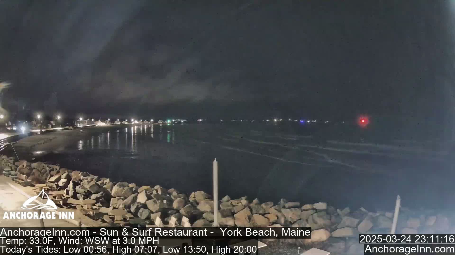 A nighttime view of a rocky shoreline and calm ocean under a dark, cloudy sky.