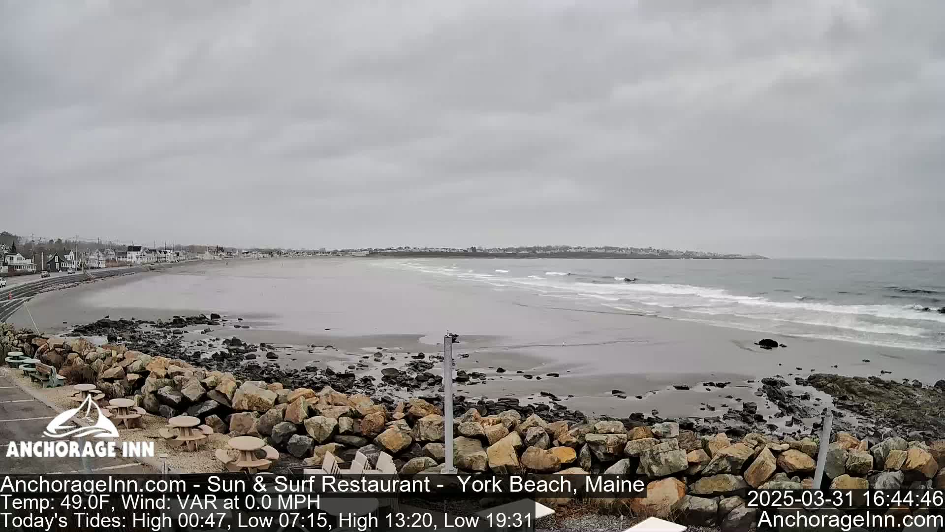 A gray, overcast day at a beach shows a wide expanse of sand and calm waves lapping the shore, with a rocky embankment in the foreground and buildings visible in the distance.
