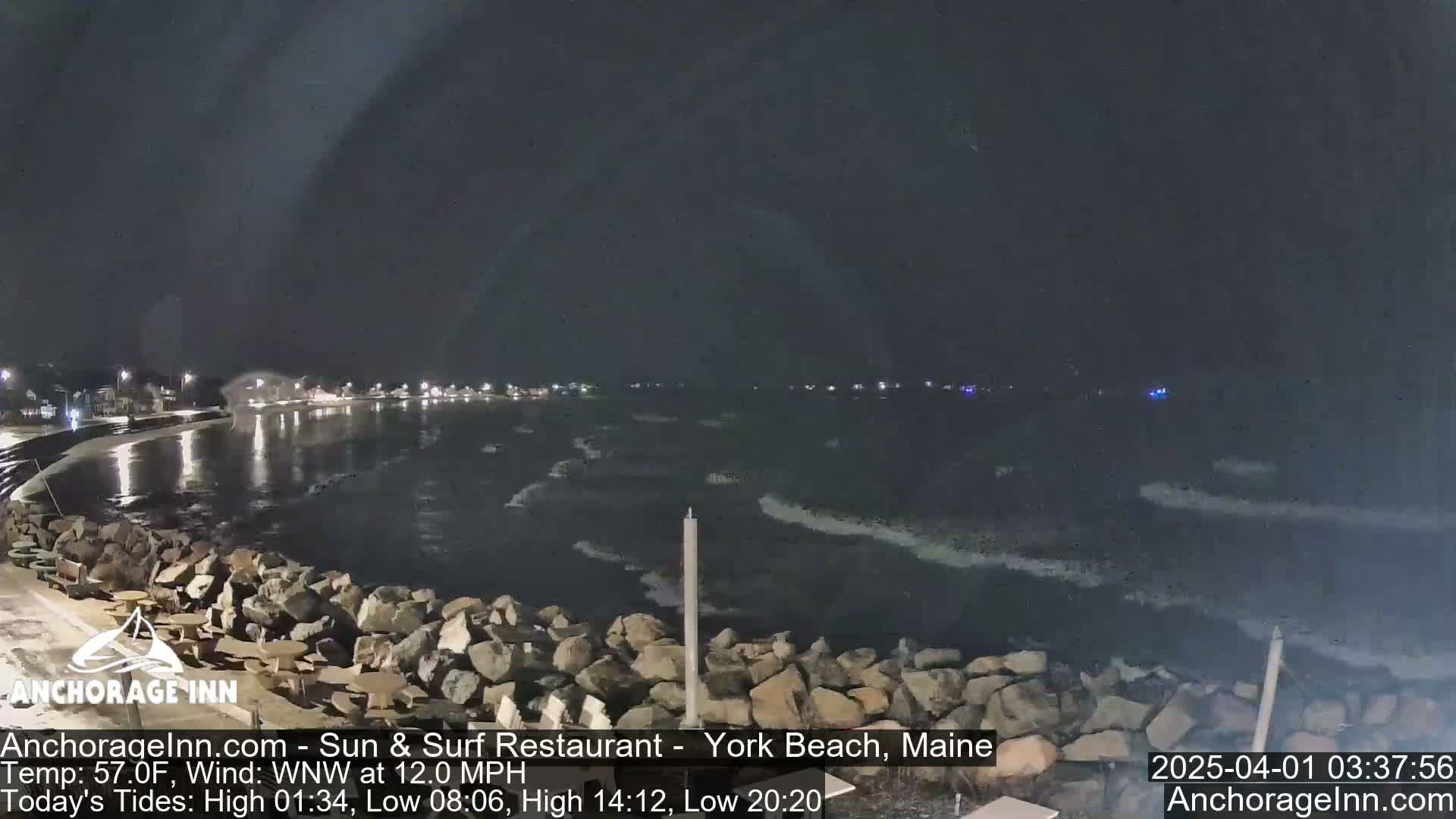 A nighttime view of a rocky shoreline with small waves lapping against it, under a dark, clear sky.