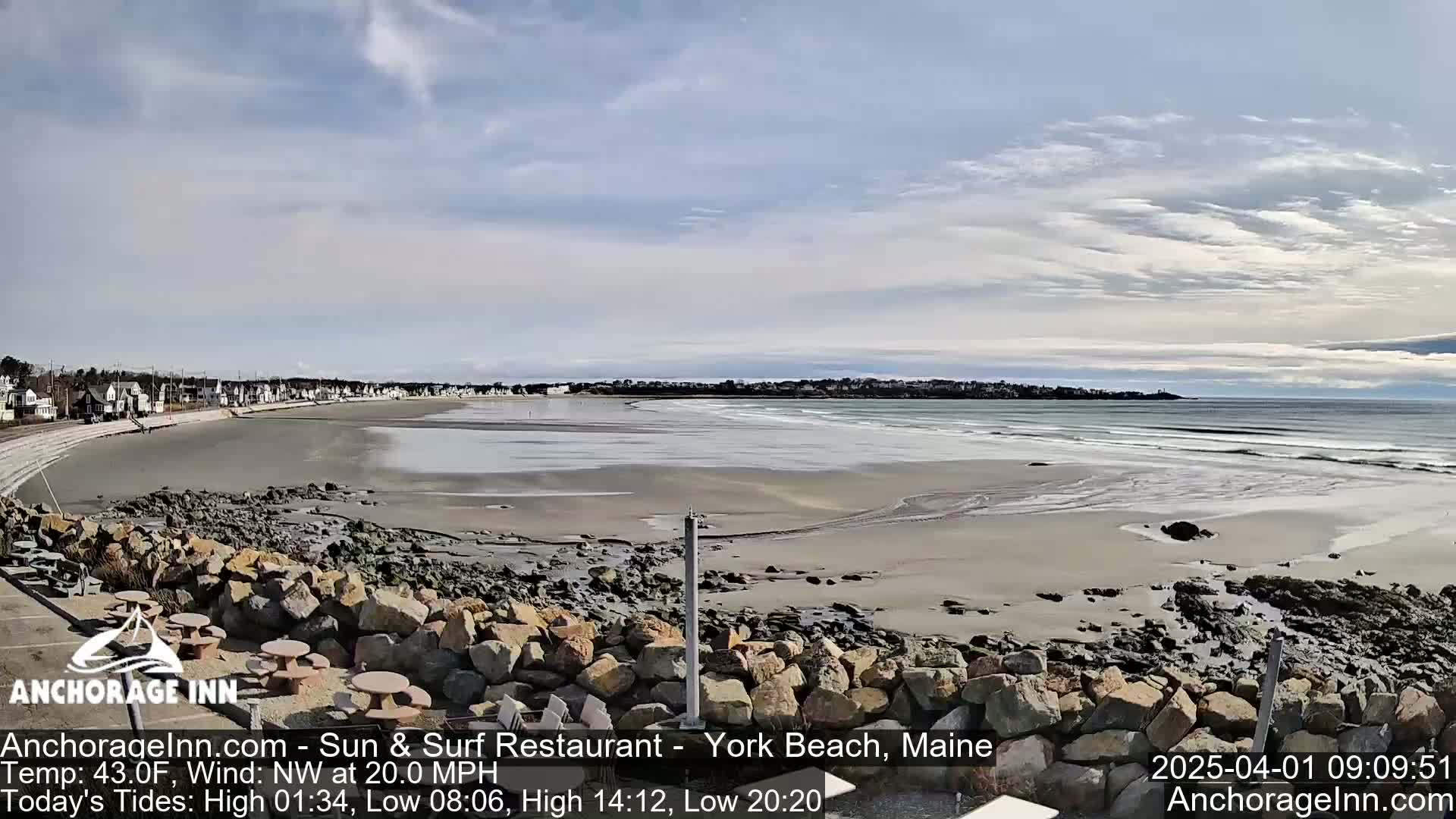 A sandy beach with small waves stretches out to a calm ocean under a mostly cloudy sky.