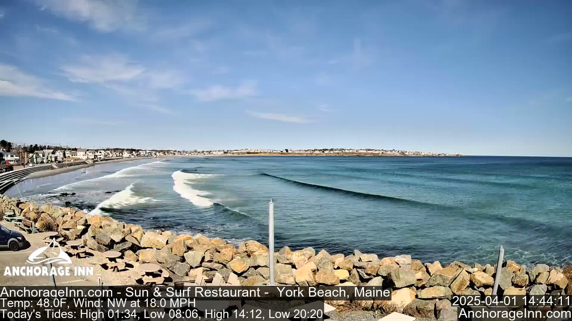 A sunny day shows a beach with gentle waves rolling onto a rocky shore, a town visible in the background.