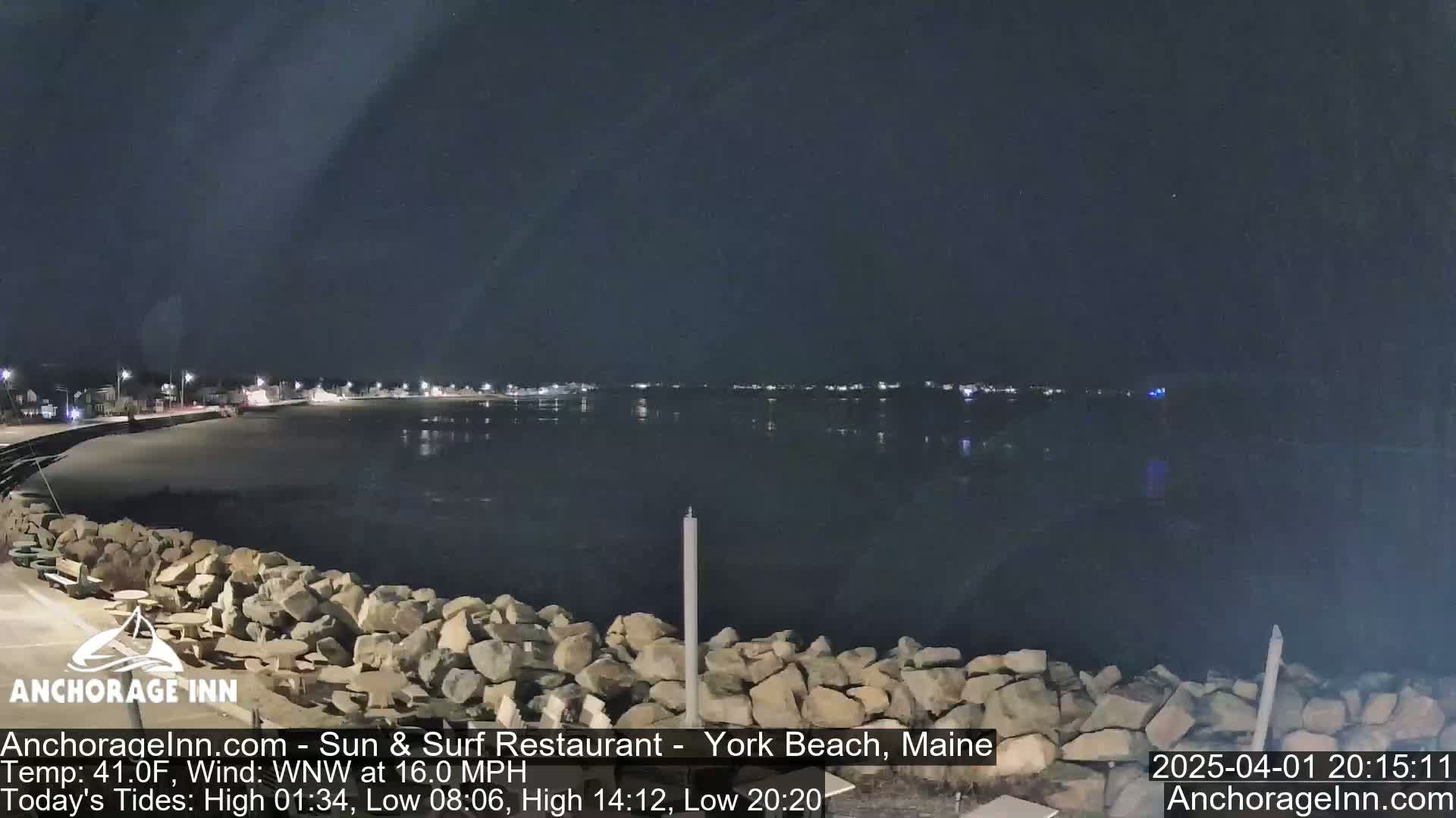 A nighttime view of a rocky shoreline and calm, dark water under a clear sky, with distant lights illuminating a town across the water.