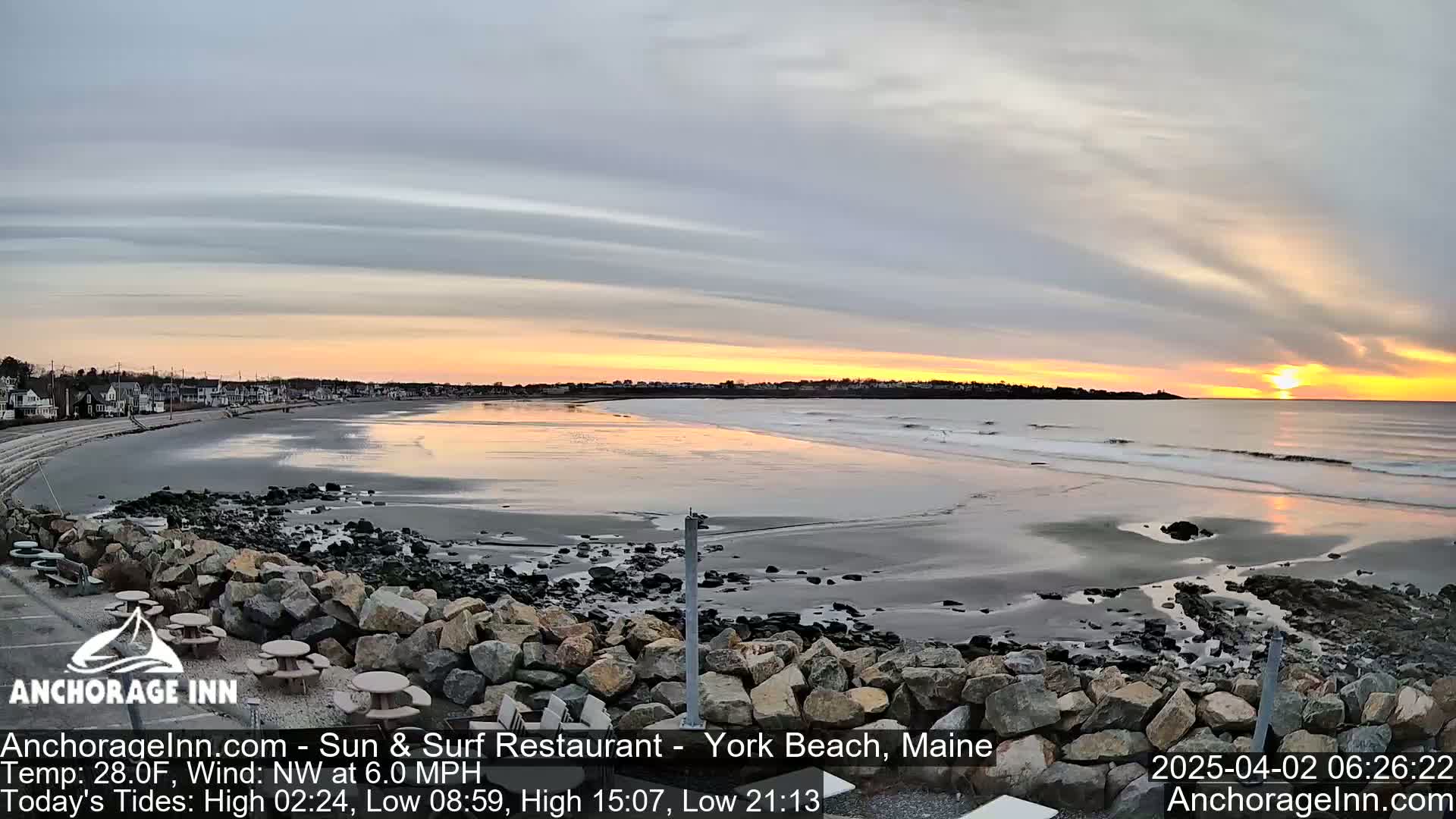 A sunrise over a calm ocean beach with a rocky shore and a row of houses in the distance, under a partly cloudy sky.