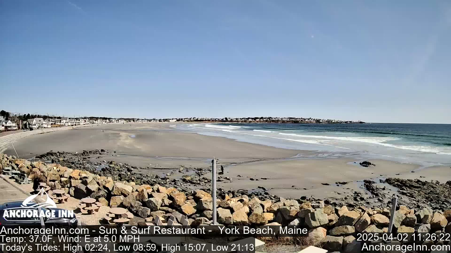 A sandy beach with gentle waves laps the shore under a clear blue sky, with a town visible in the distance.