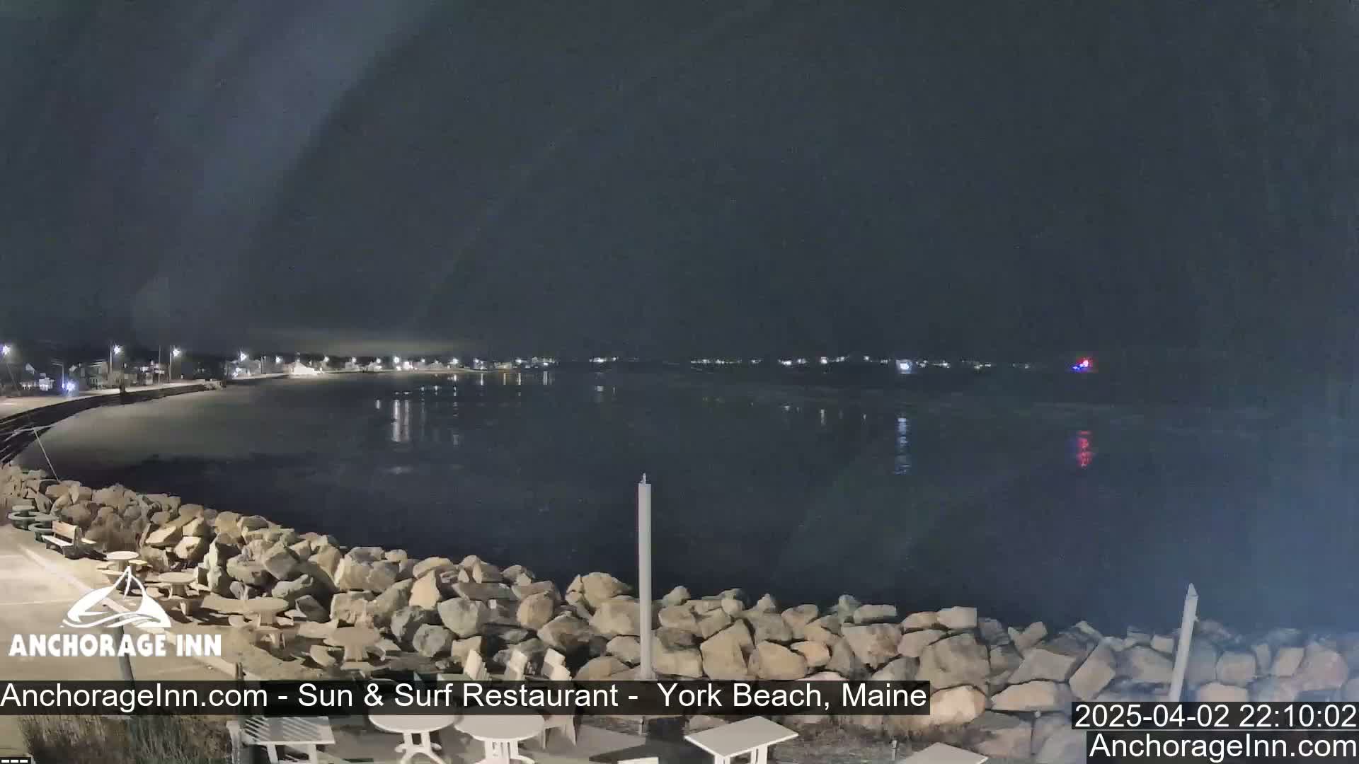 A nighttime view of a calm, dark body of water next to a rocky shoreline with lights visible in the distance under a dark sky.