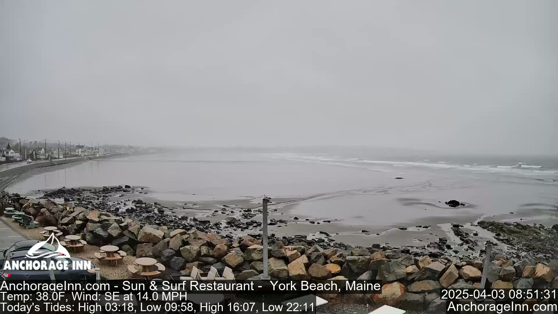 A foggy, overcast beach scene shows a rocky shoreline with a calm ocean and a distant line of buildings.