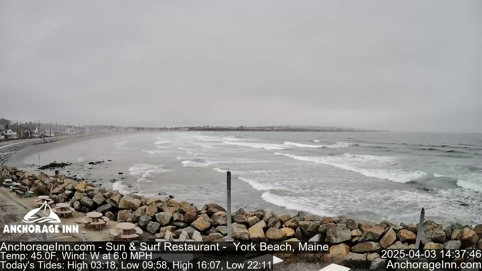 A rocky shoreline with picnic tables overlooks a gray, overcast ocean with gentle waves.