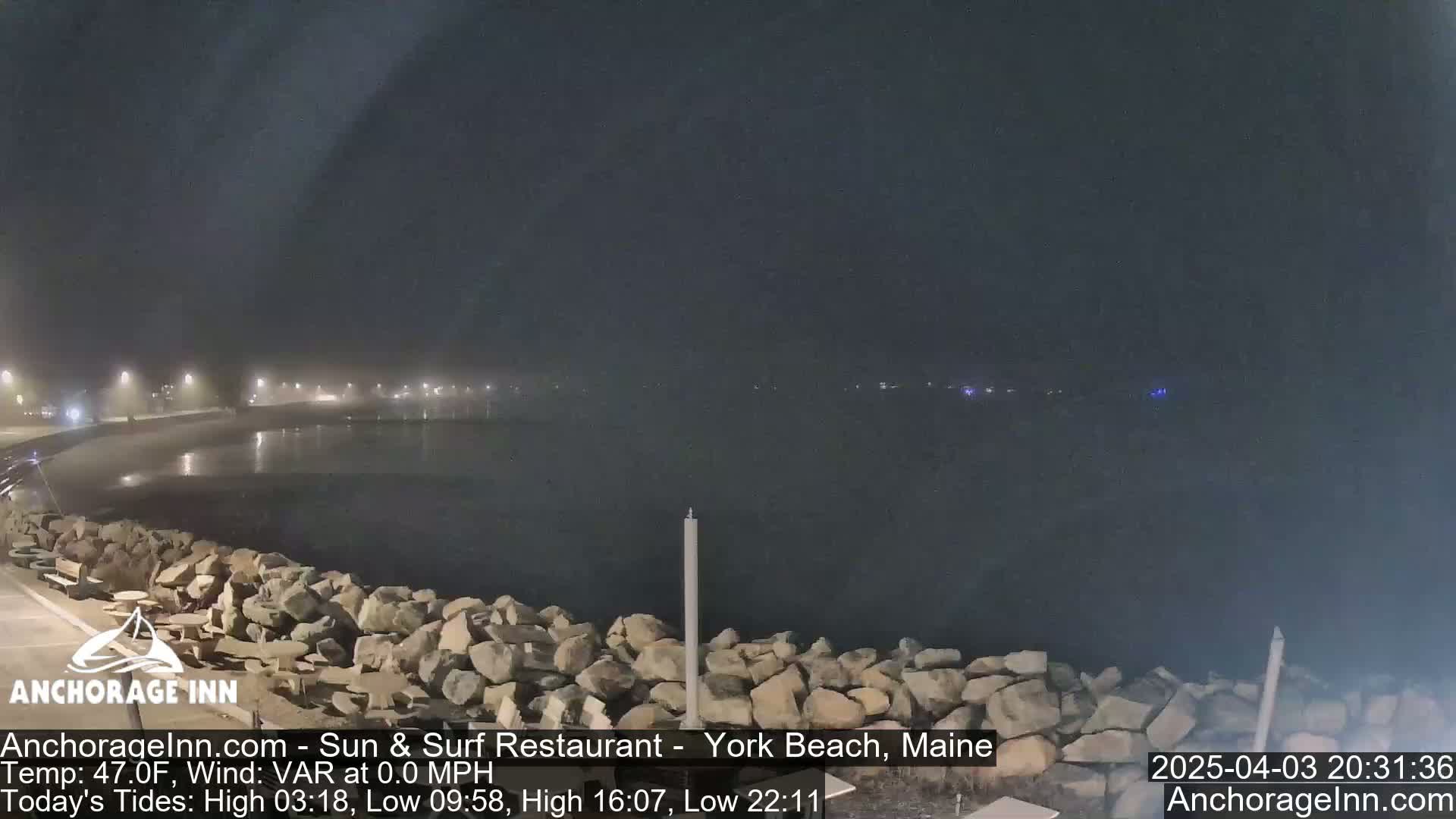 A nighttime, foggy view of a rocky shoreline and calm water, with distant lights visible across the water.