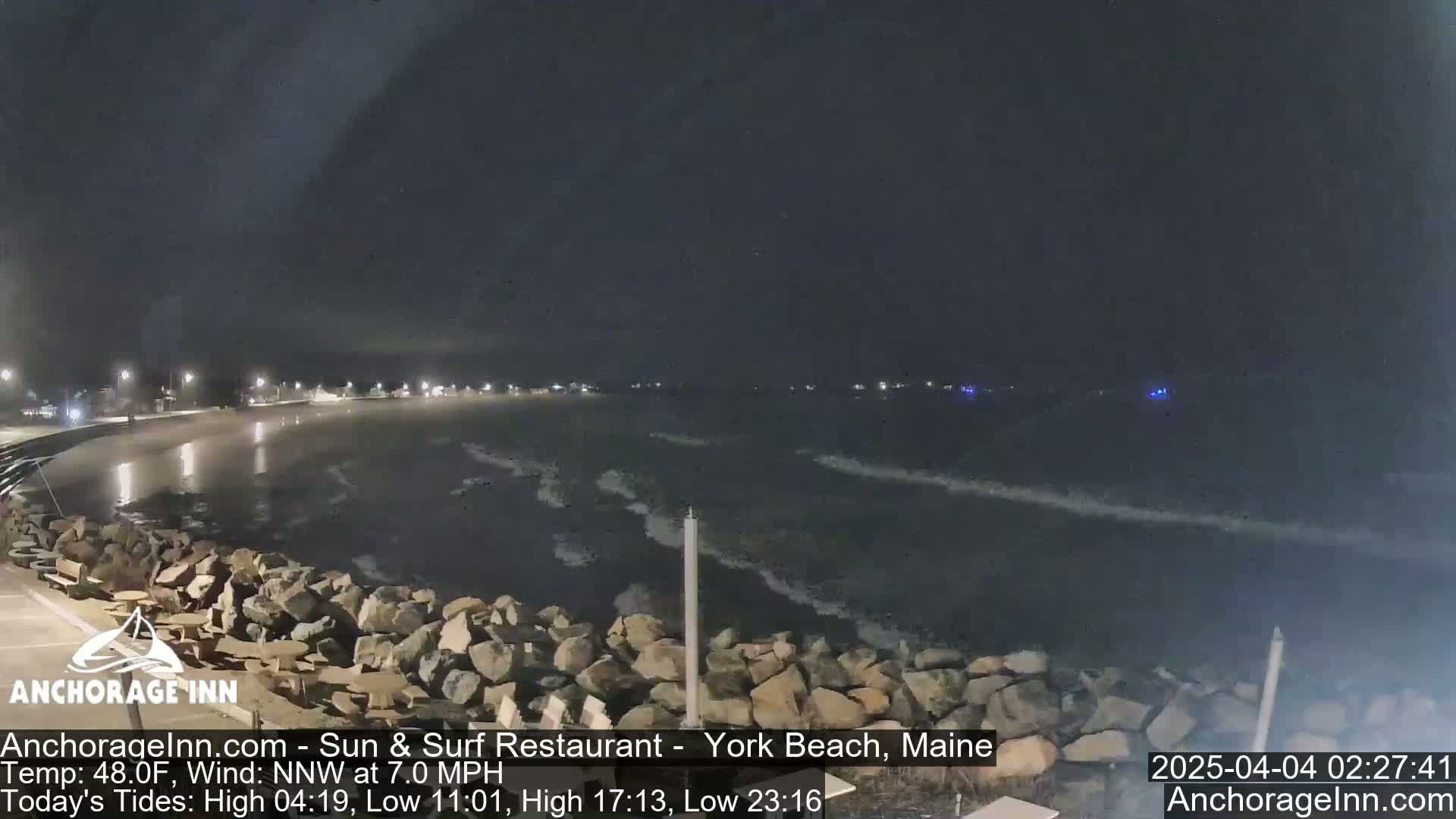A nighttime view of a rocky shoreline and dark ocean with small waves under a cloudy sky.