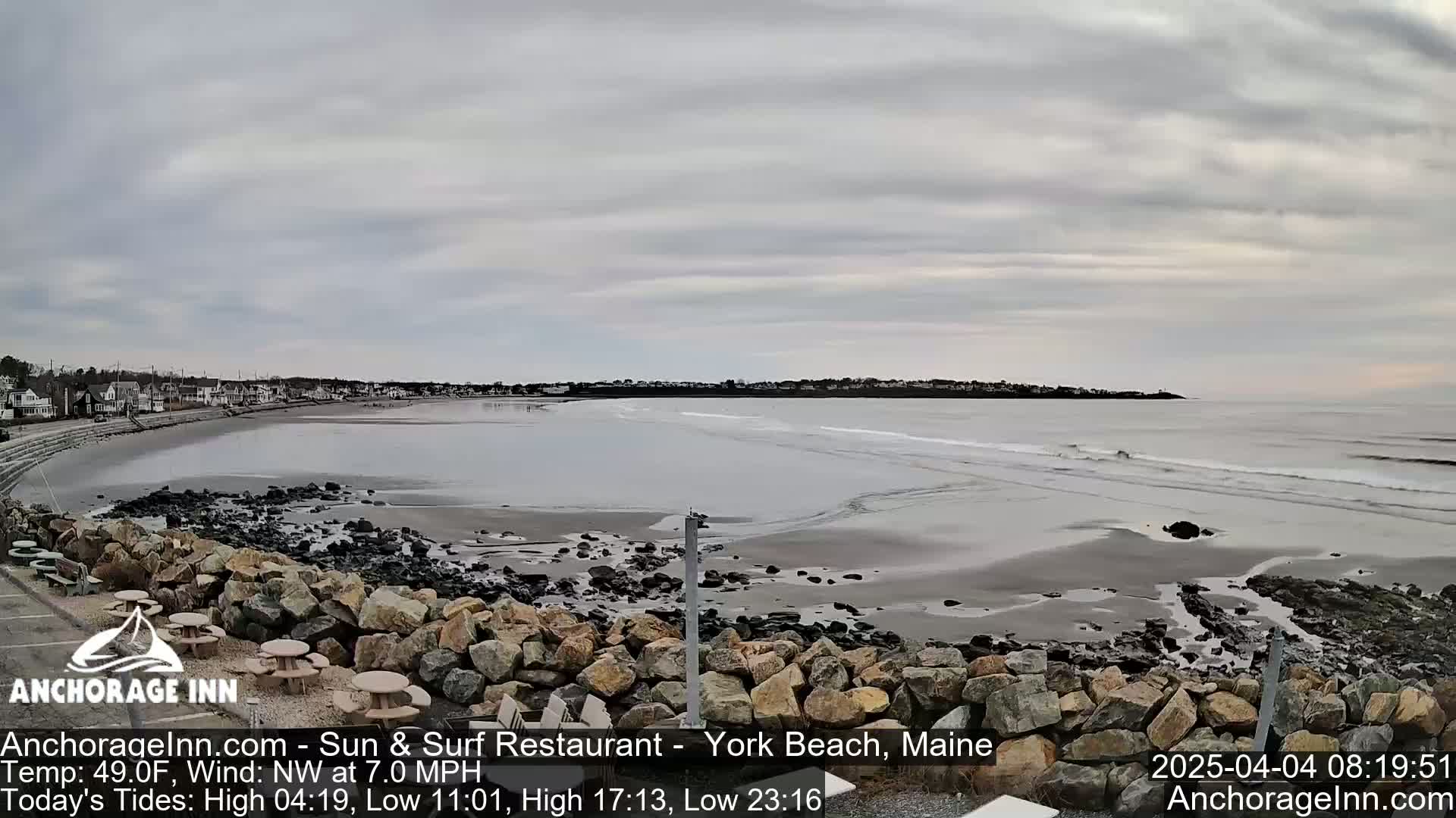 A wide, sandy beach with small waves under an overcast sky is seen from a rocky overlook, with a town visible in the distance.