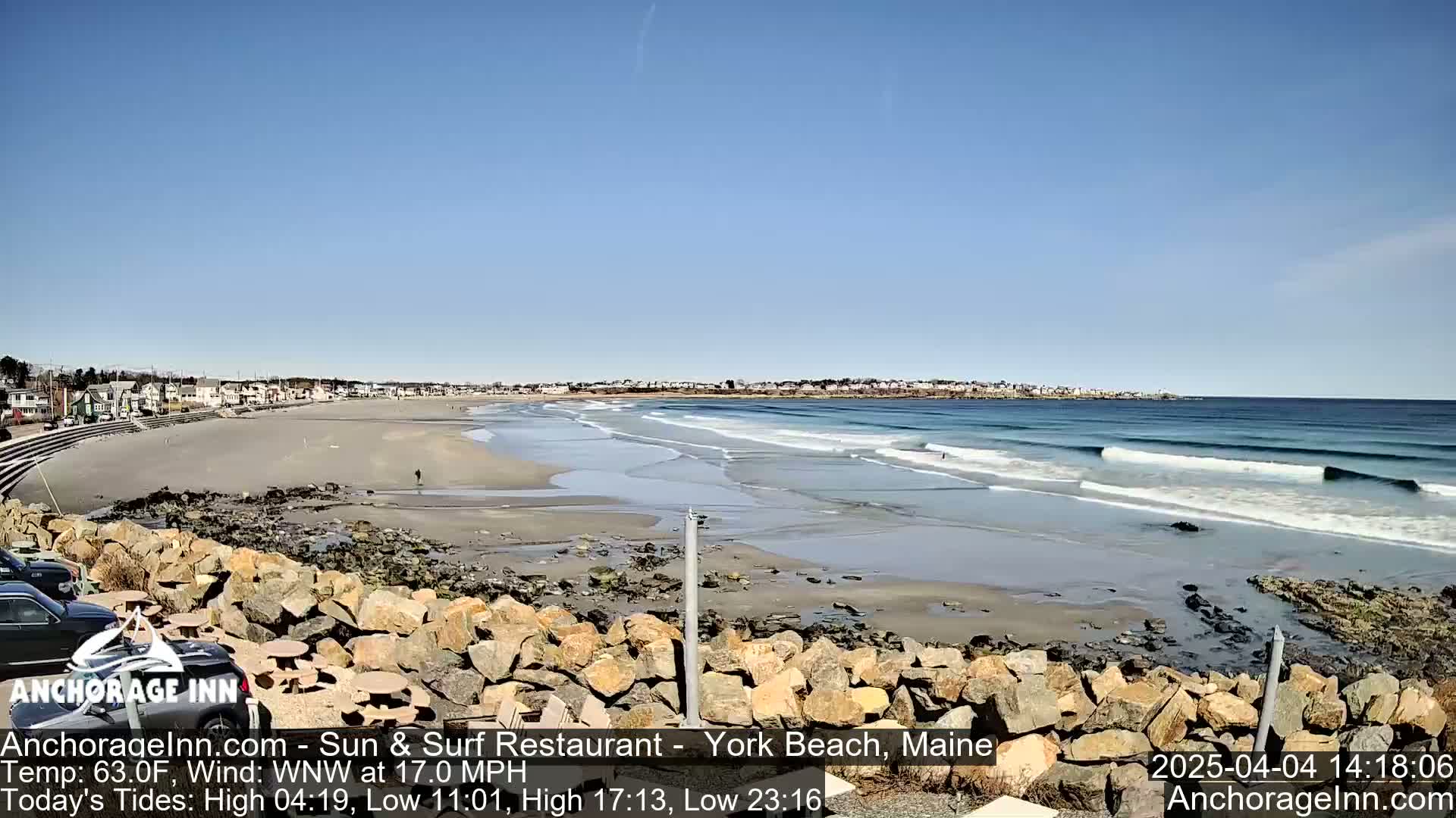 A sandy beach with gentle waves under a clear blue sky is viewed from a rocky outcrop, with houses visible in the distance.