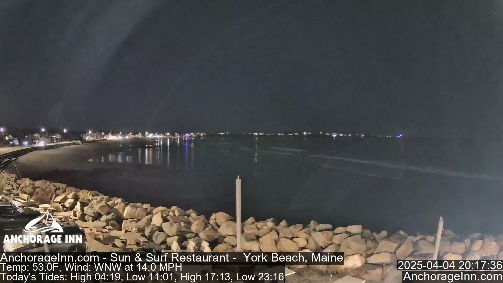 A nighttime view of a calm, dark ocean with a rocky shoreline and distant lights from a town under a clear, dark sky.