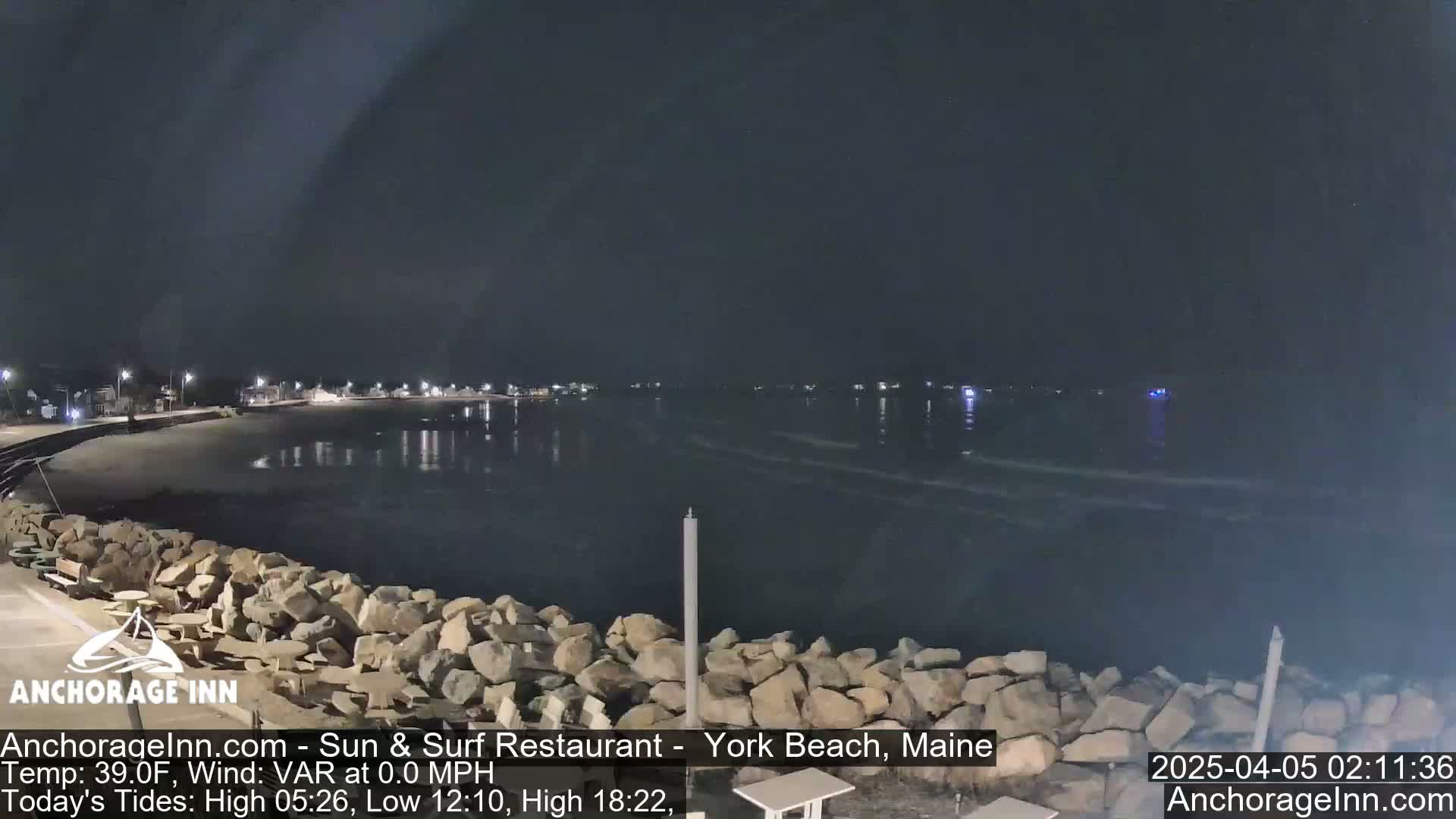 A dark nighttime scene shows a calm body of water lapping gently against a rocky shoreline, with lights visible in the distance along a beach.