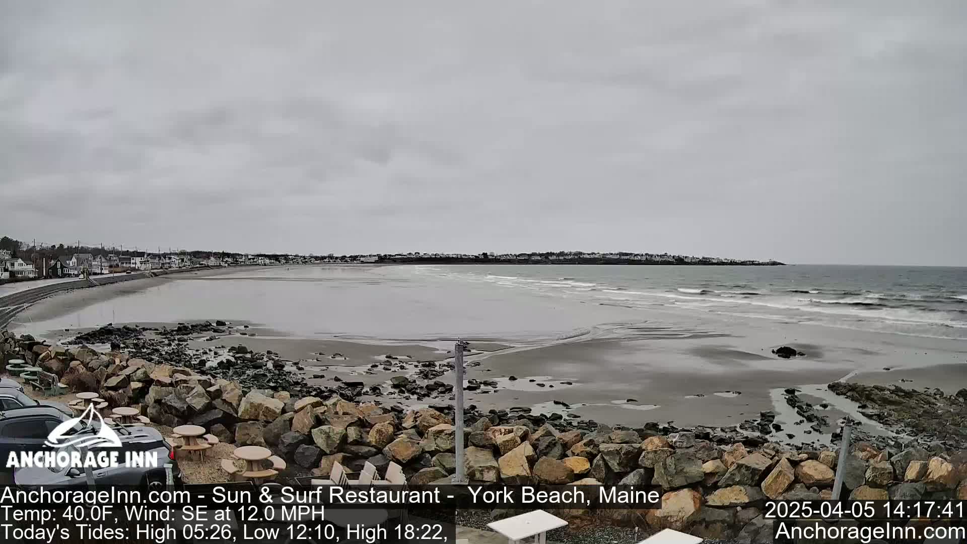 A gray, overcast day shows a sandy beach with gentle waves lapping the shore, backed by a rocky embankment and a line of buildings in the distance.