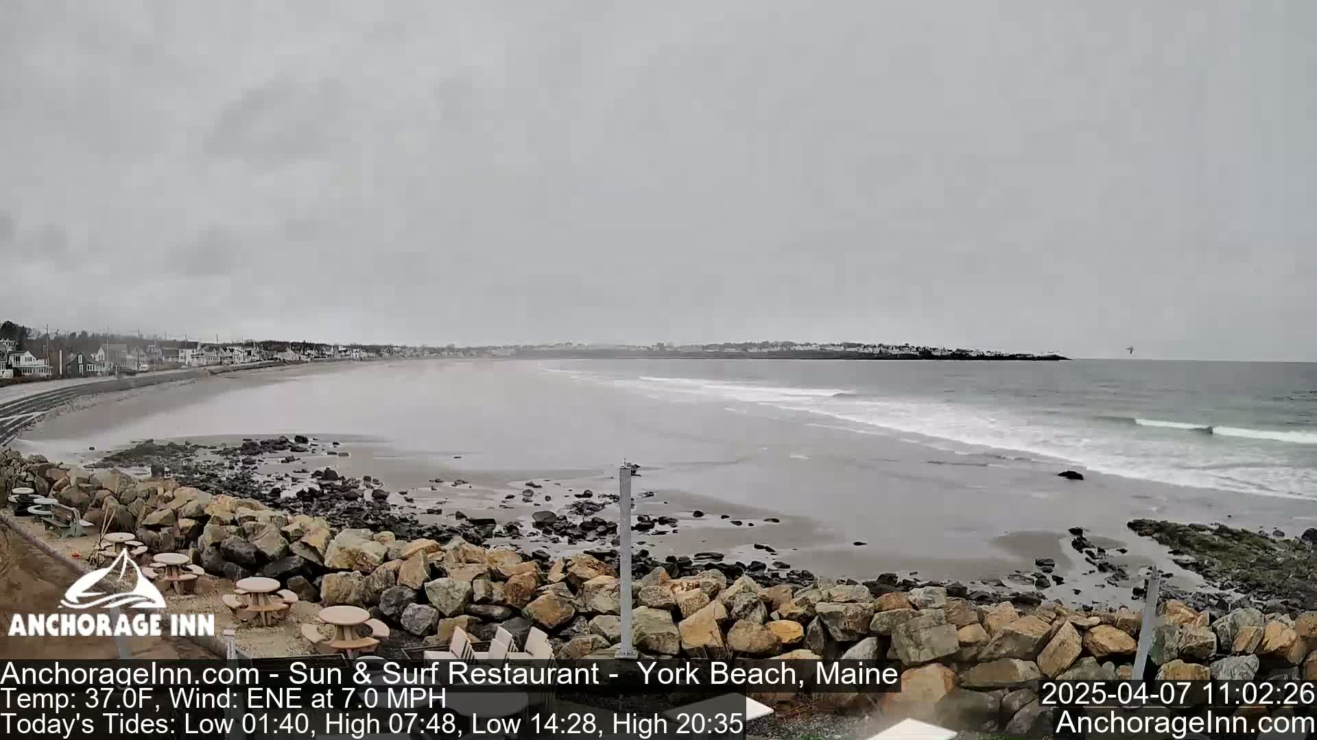A wide, sandy beach with gentle waves under an overcast sky is viewed from a rocky outcrop, with buildings visible in the distance.