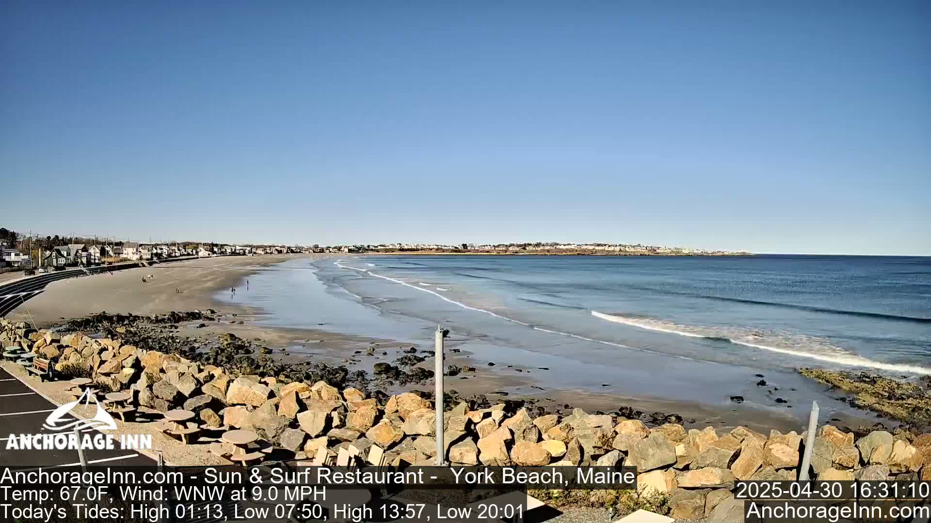 A sandy beach with gentle waves stretches out to a calm ocean under a clear blue sky, backed by a line of buildings.