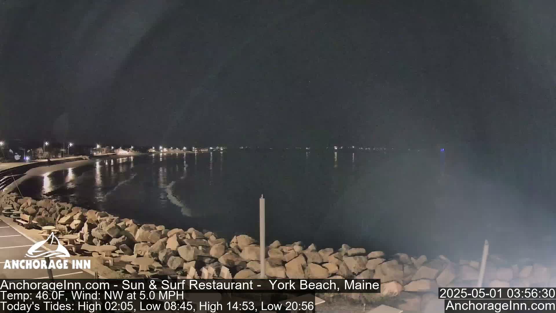 A nighttime view of a calm body of water, with a rocky shoreline and distant lights visible across the water under a dark sky.