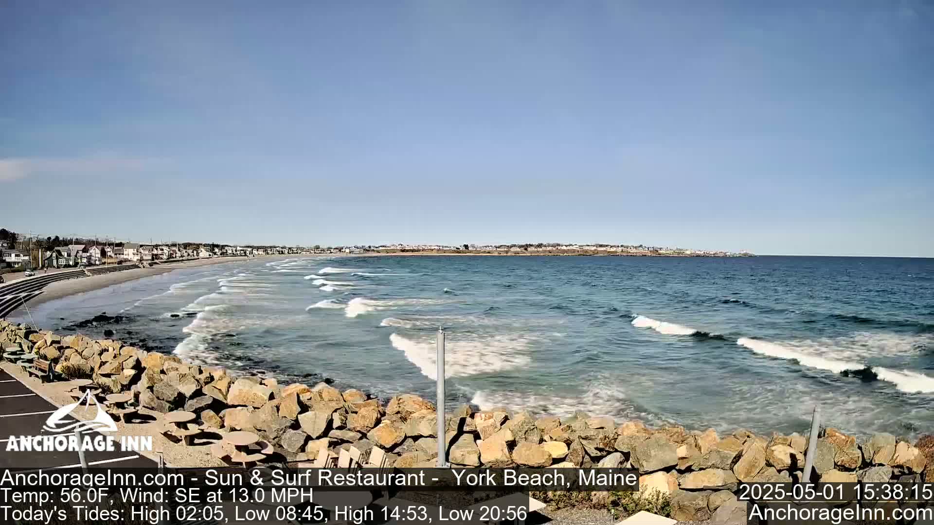 A sandy beach with gentle waves rolls onto the shore, backed by a town under a clear blue sky.