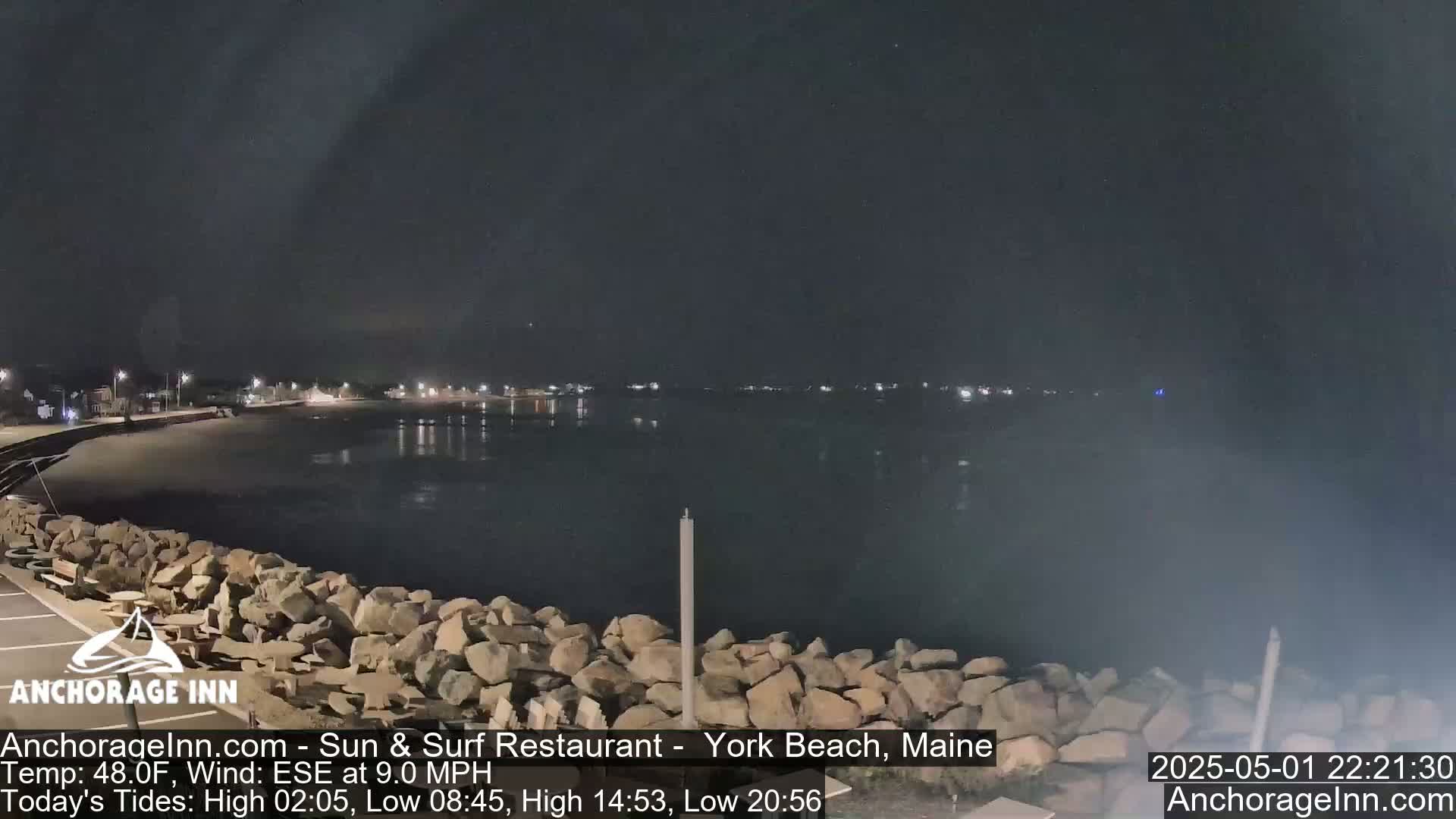 A nighttime view of a calm body of water with a rocky shoreline and faintly lit buildings in the distance.