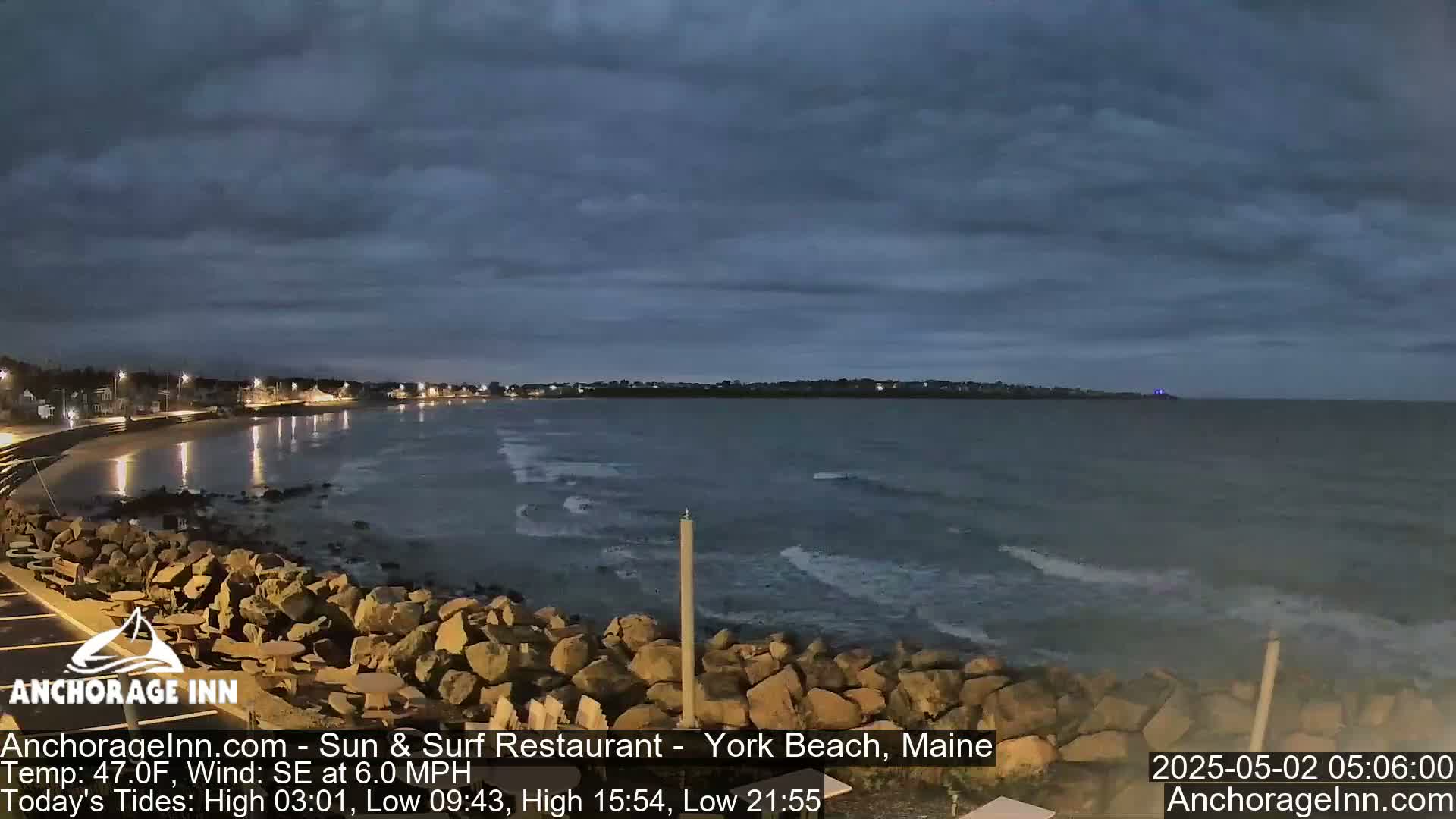 A dark, overcast evening scene shows a rocky shoreline with gentle waves lapping the coast, and a town visible in the distance.