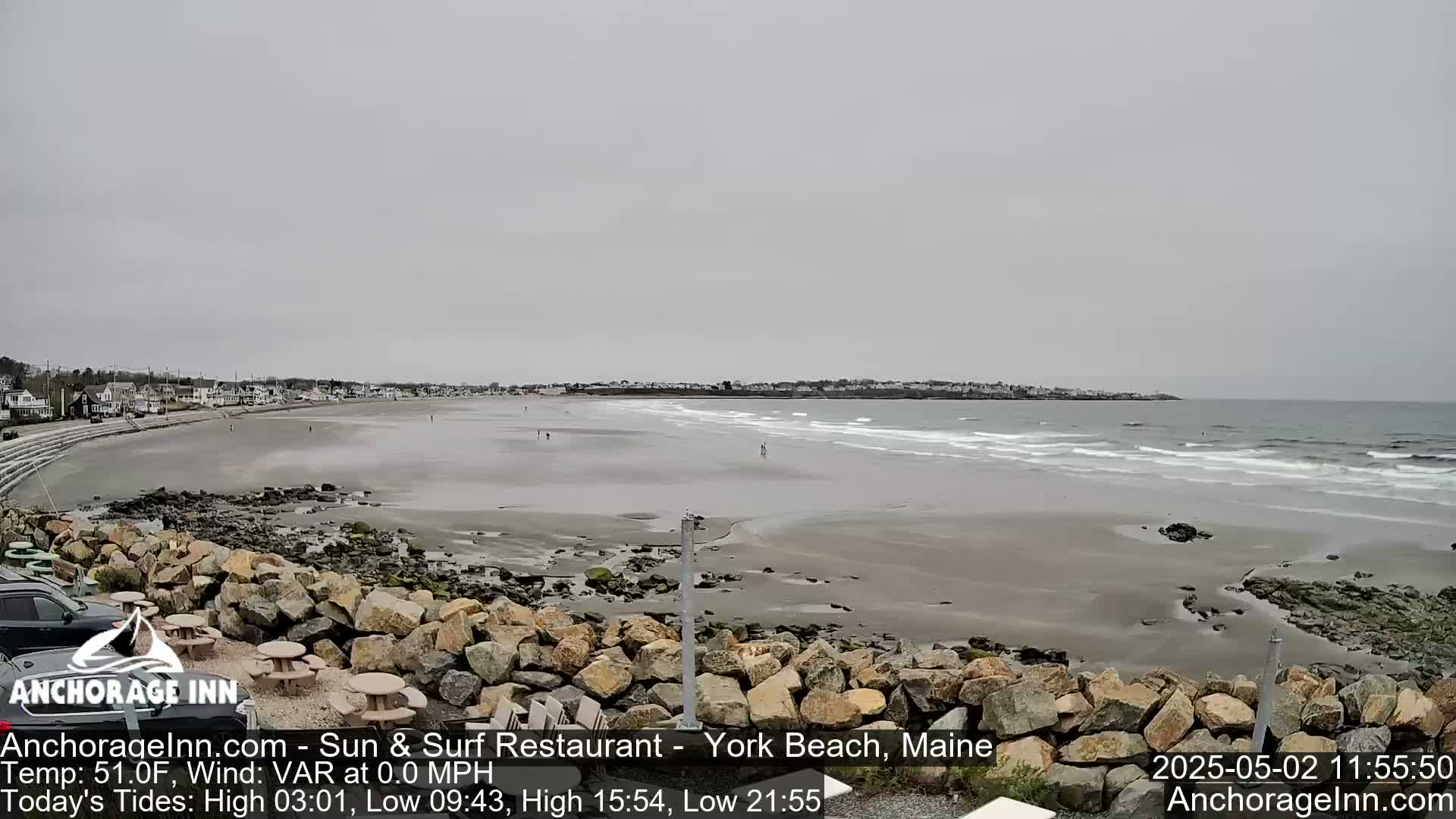 A wide, sandy beach stretches toward a calm ocean under an overcast sky, with a rocky shoreline in the foreground and buildings visible in the distance.