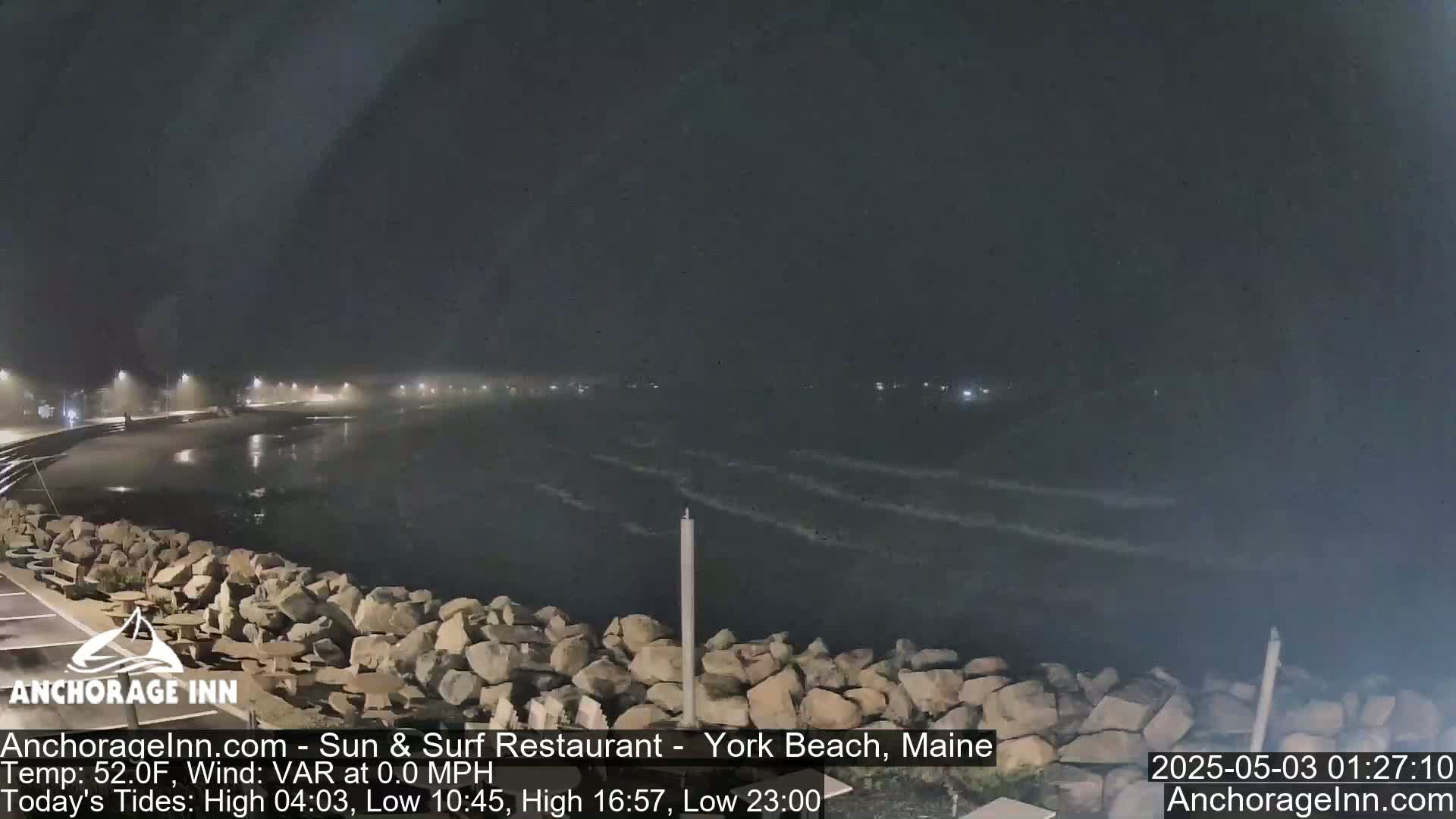 A nighttime view of a rocky shoreline and dark, gently waving ocean under a hazy sky.