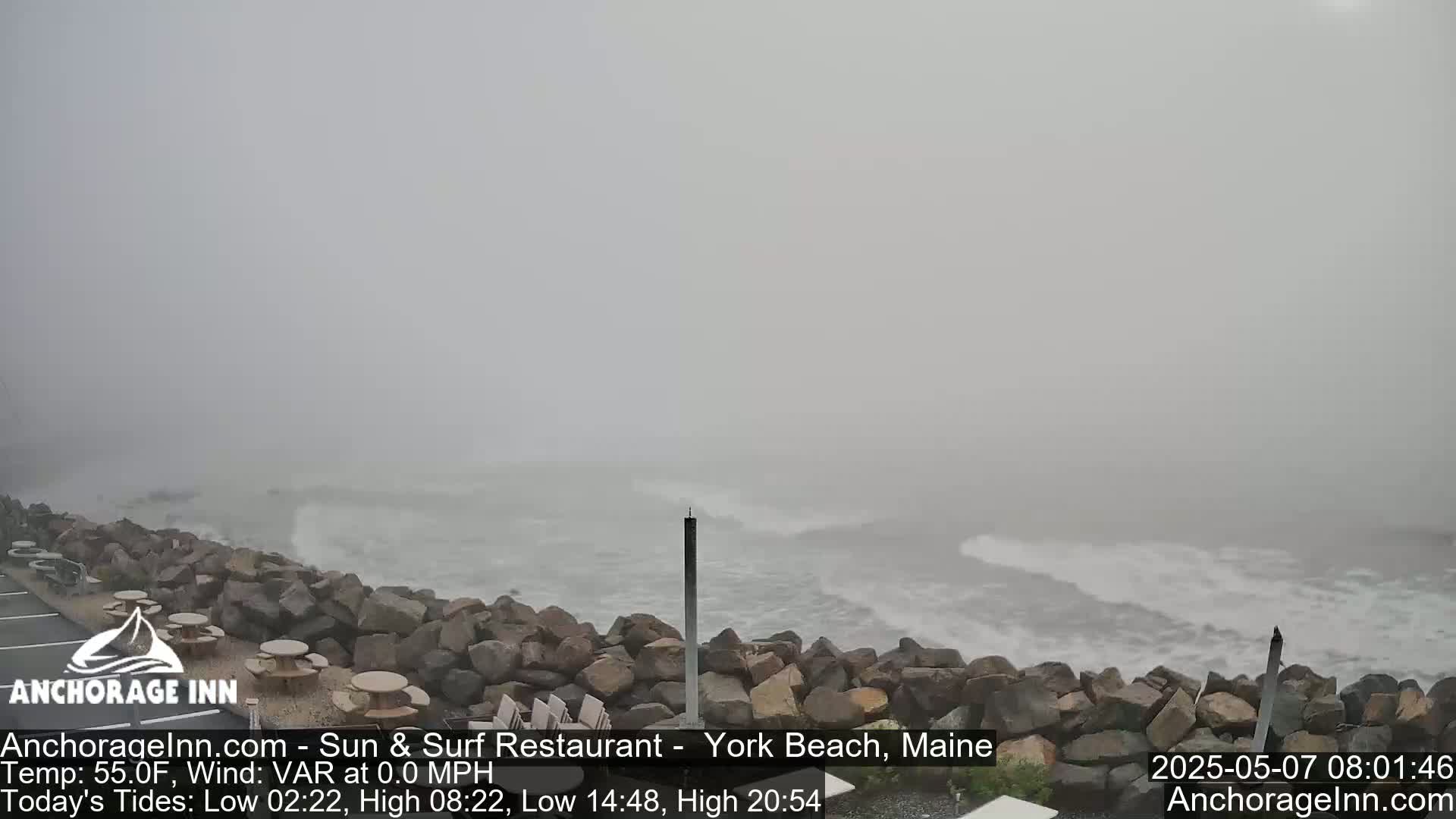 A foggy, overcast coastal scene shows a rocky shoreline with small waves breaking against it.
