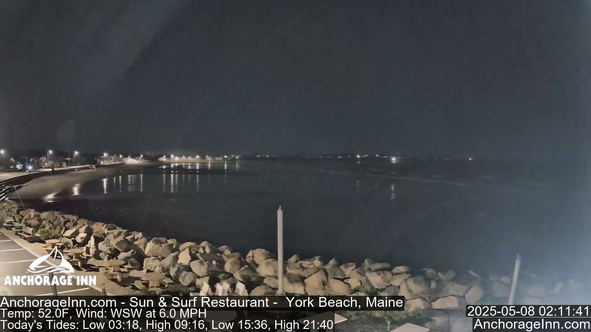 A nighttime view of a calm, dark body of water with a rocky shoreline and dimly lit buildings in the distance under a clear sky.