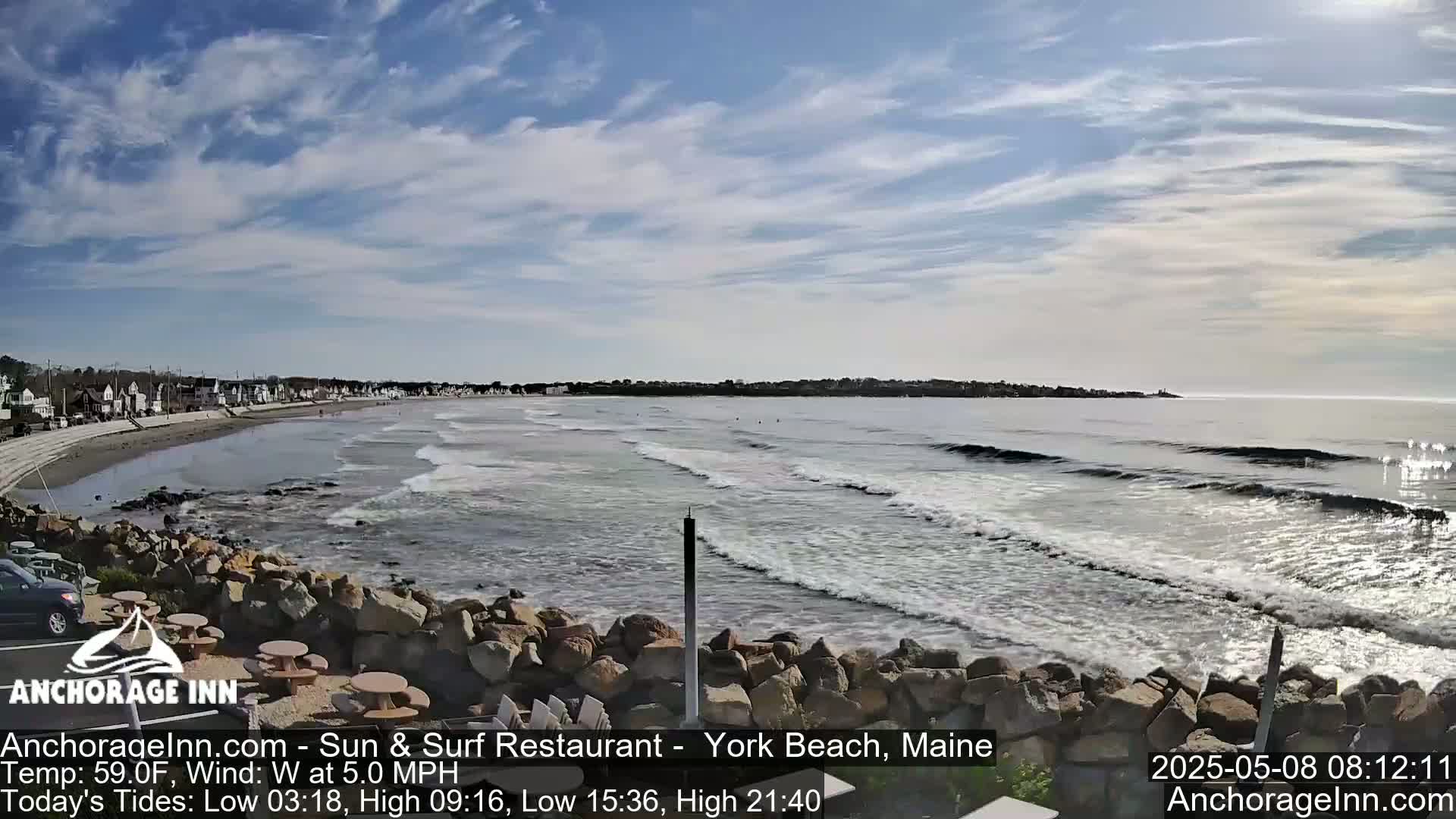 A mostly sunny day shows a beach with gentle waves lapping a rocky shore, and a row of houses visible in the distance.