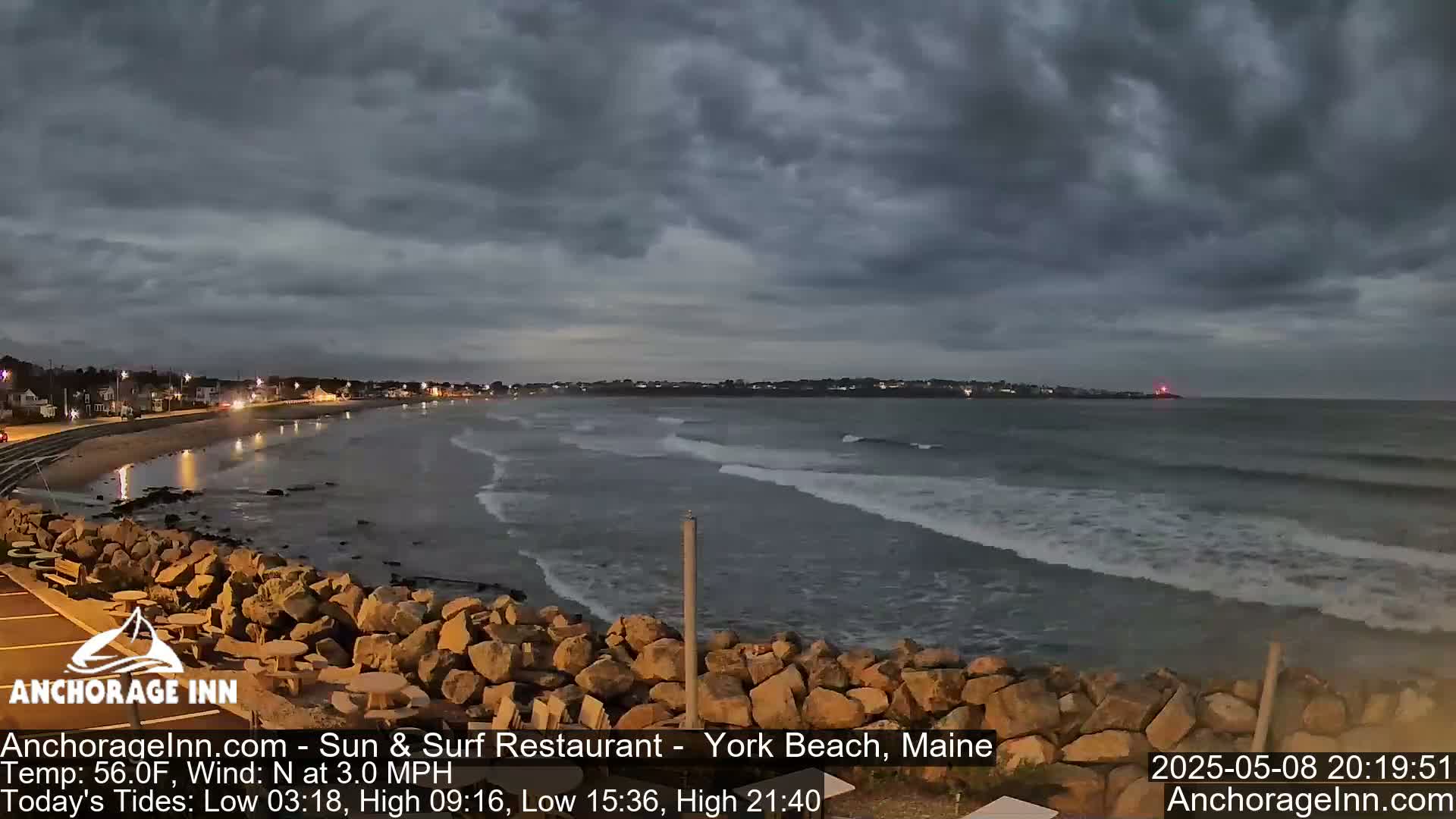 A mostly cloudy evening scene shows a rocky shoreline and calm ocean waves lapping gently against the shore, with a few lights visible in the distance.