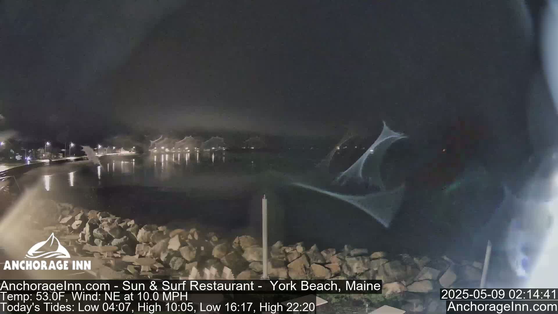 A nighttime view of a calm body of water, partially obscured by what appears to be spray or mist, with a rocky shoreline and dimly lit buildings in the distance under a dark sky.