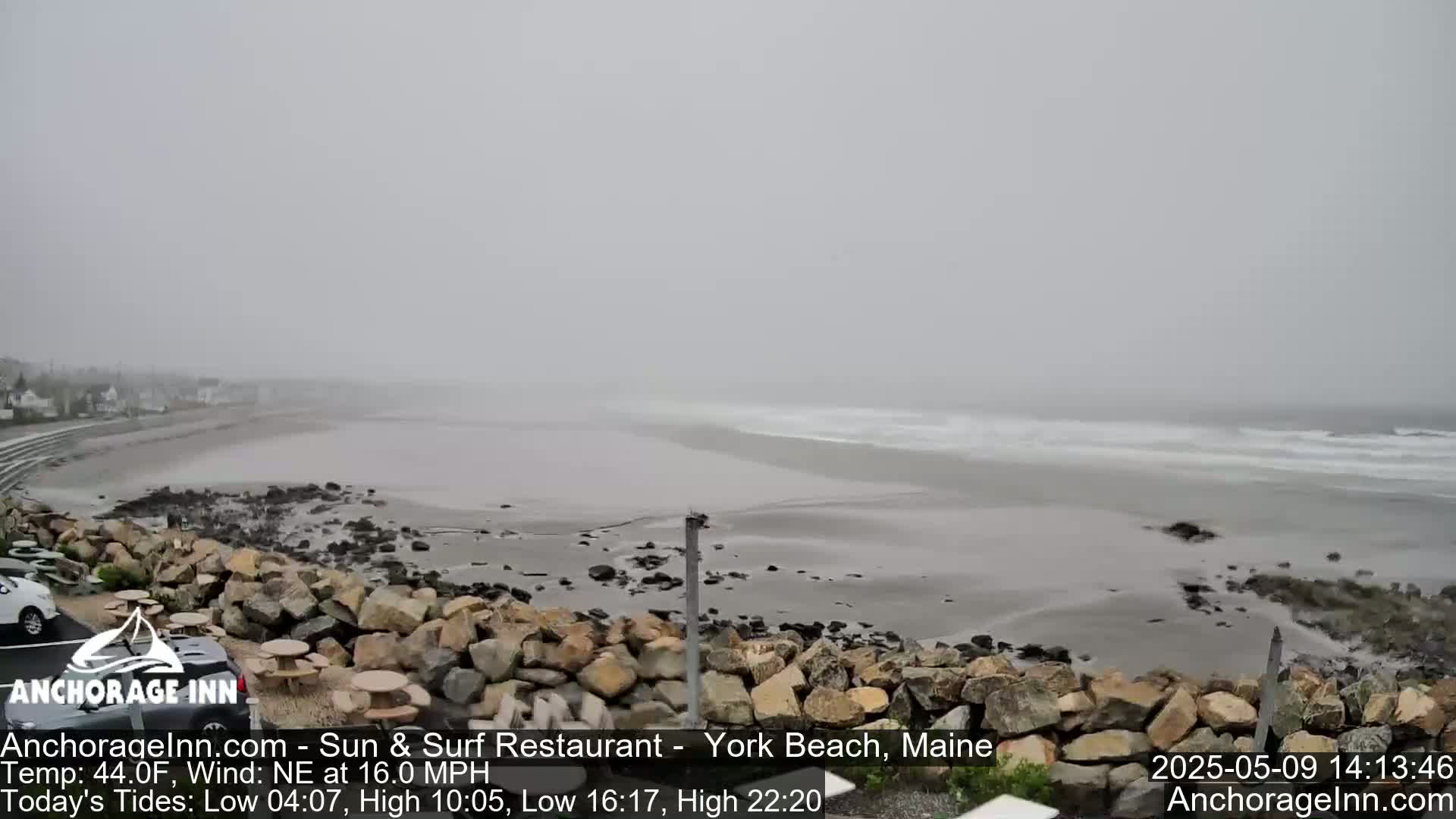 A foggy beach scene shows a gray ocean with small waves breaking on a sandy shore, behind a rocky embankment and a parking lot with cars.
