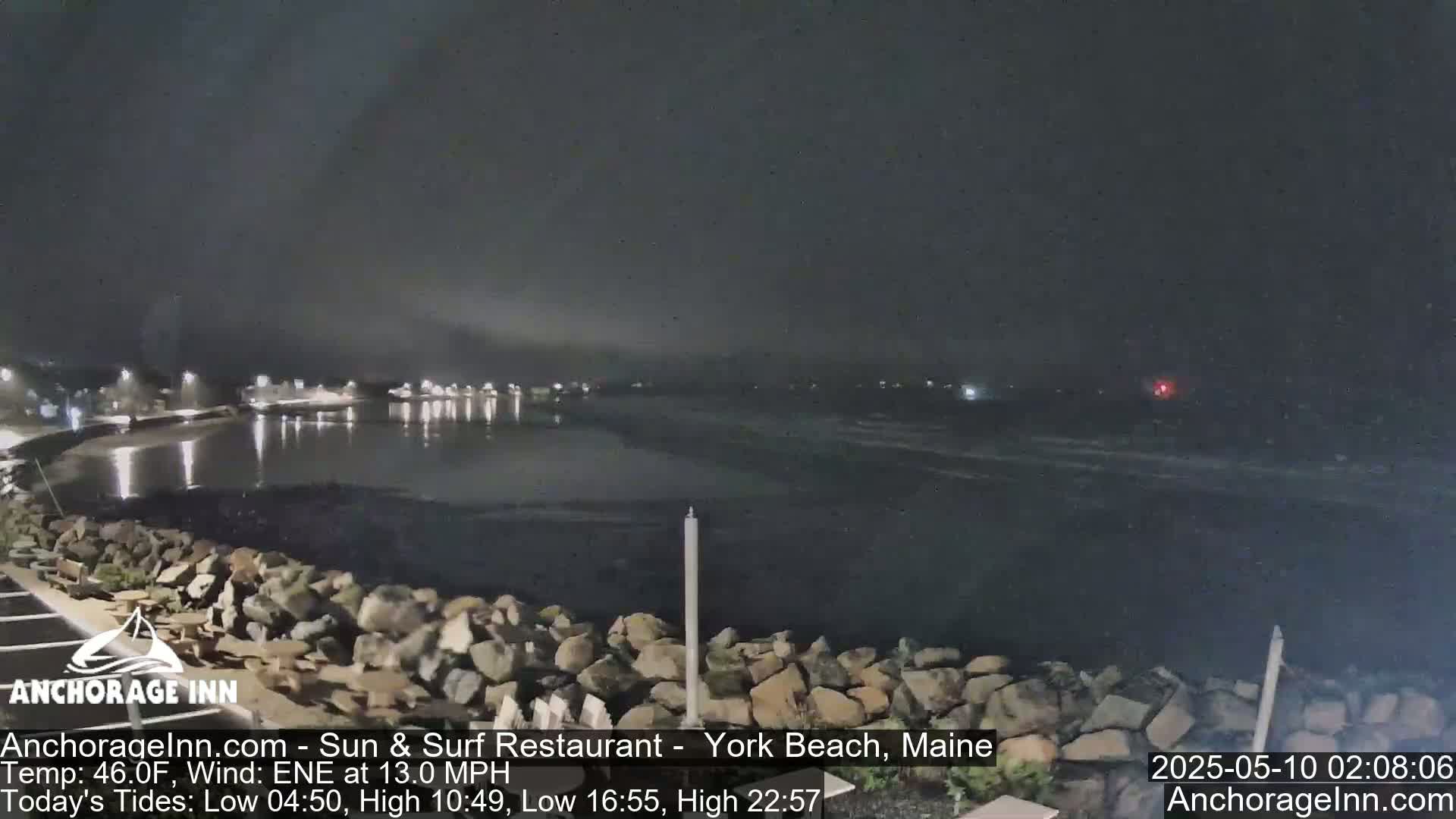 A nighttime view of a dark, rocky shoreline and calm water, with lights visible in the distance.