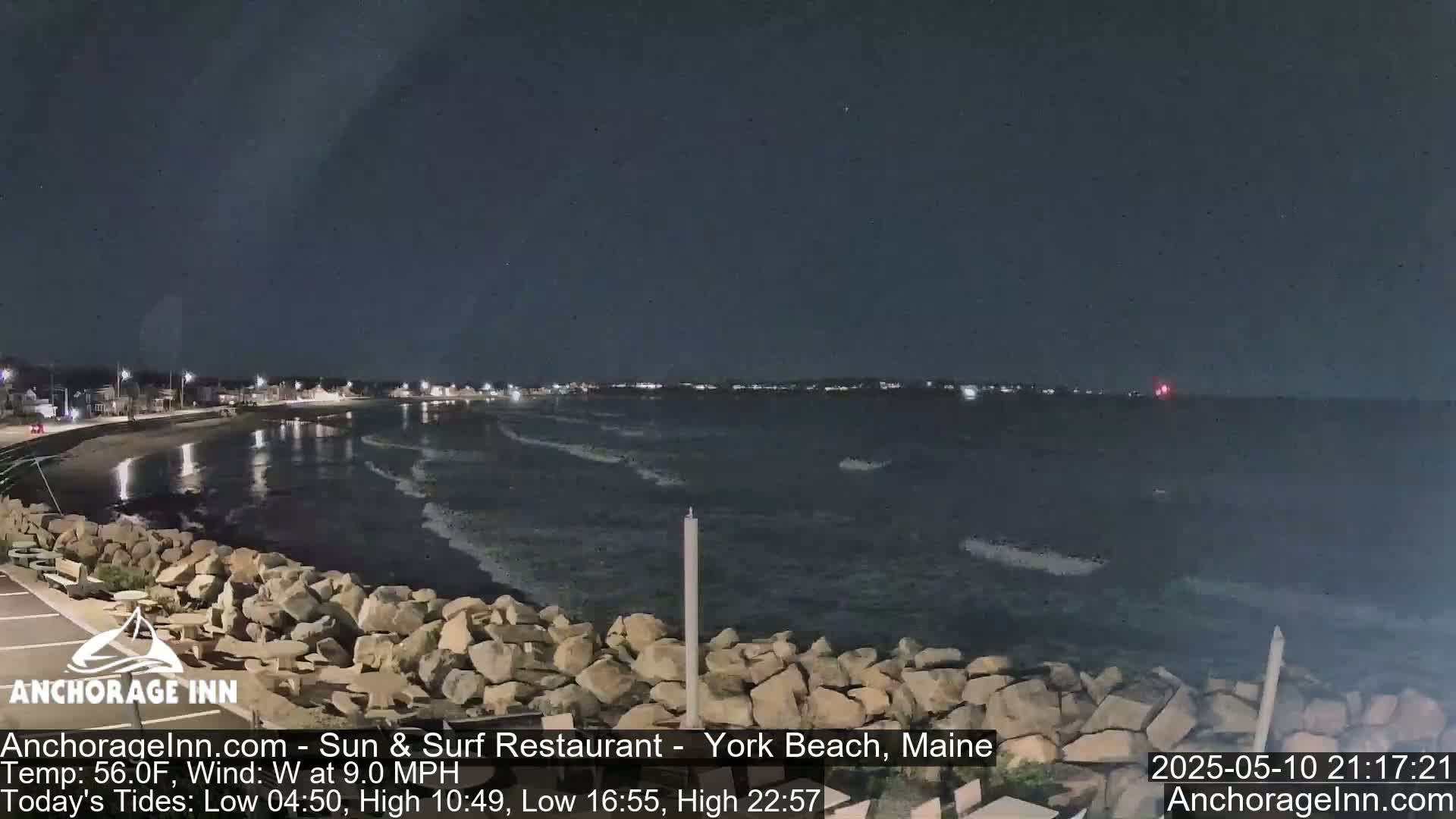 A nighttime view of a rocky shoreline and calm ocean with a town faintly lit in the distance under a dark, clear sky.