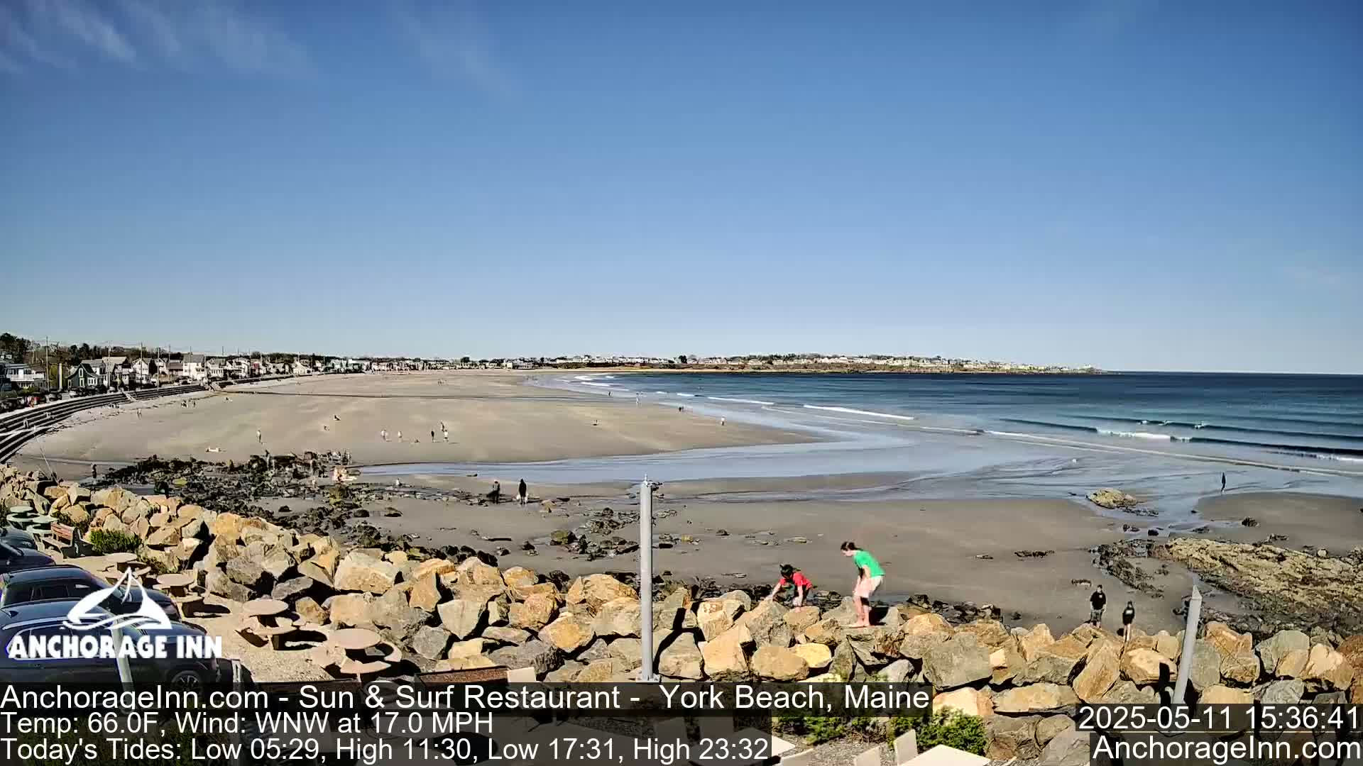 A sandy beach with a few people and gentle waves under a clear blue sky is viewed from a rocky overlook.