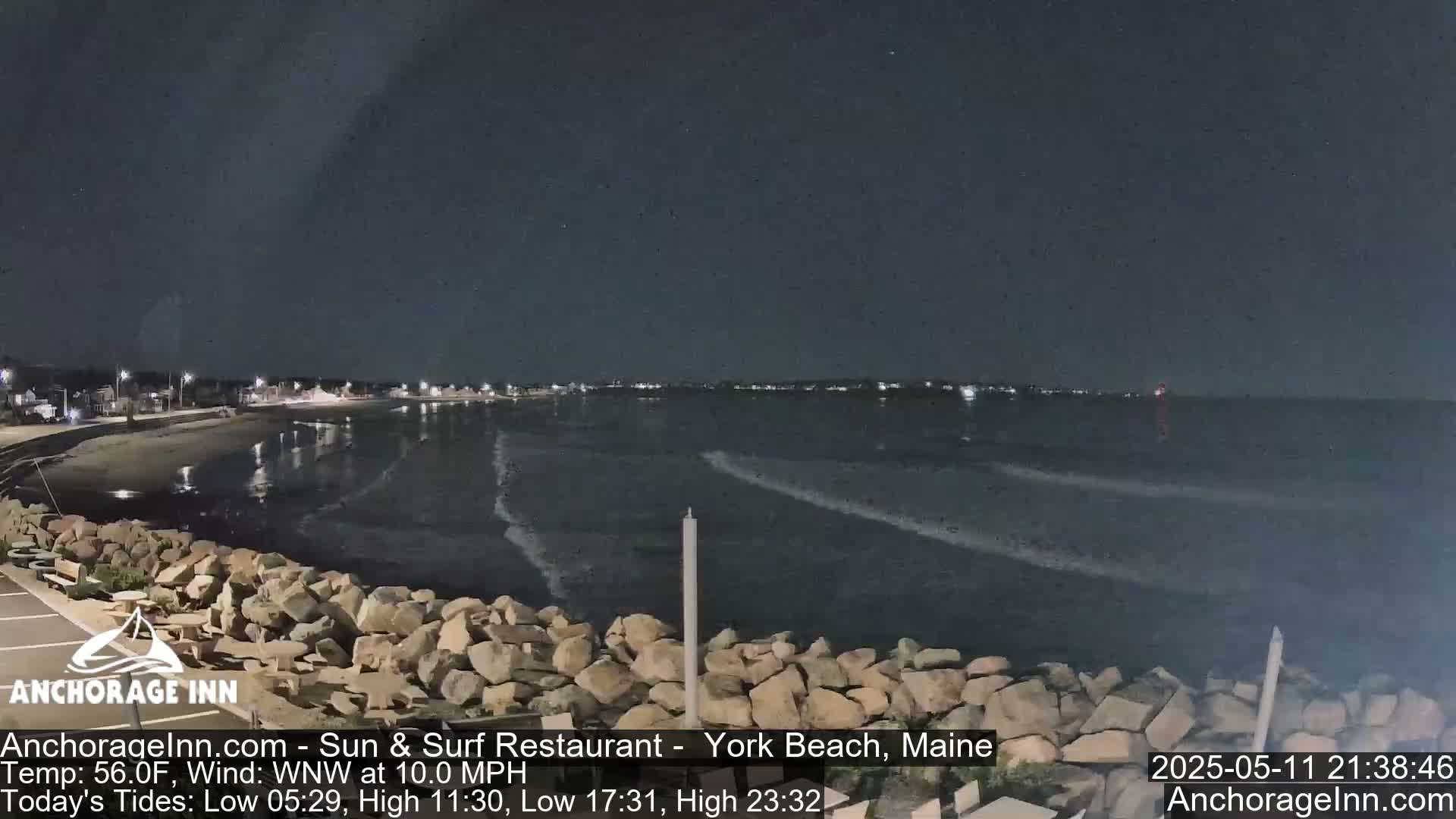 A nighttime view of a rocky shoreline and calm ocean with distant lights on the horizon under a dark sky.
